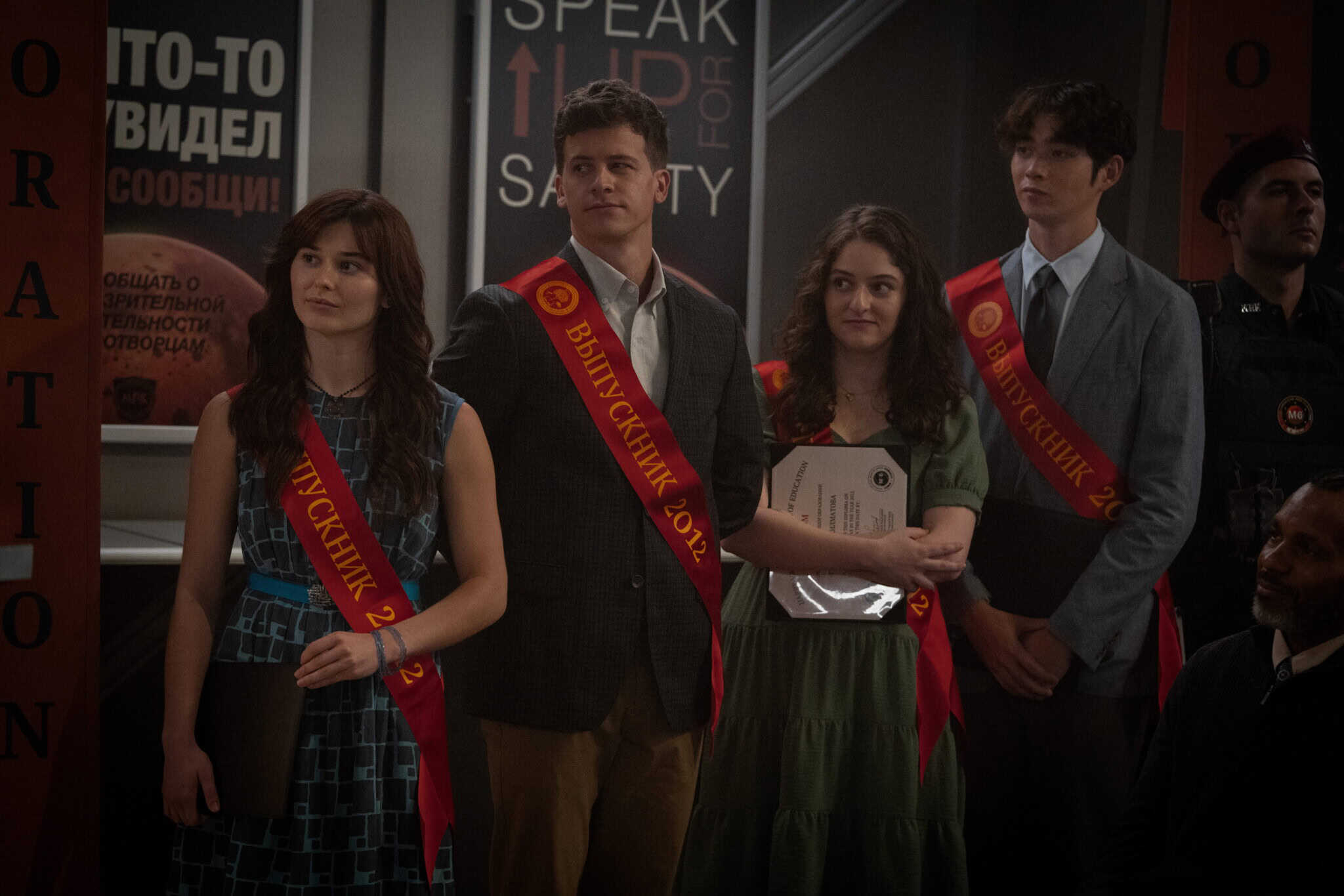 Four young people wearing red sashes stand together at a ceremony. One holds a certificate. They are dressed formally and appear to be graduating. A security officer and signs with Russian text are visible in the background.