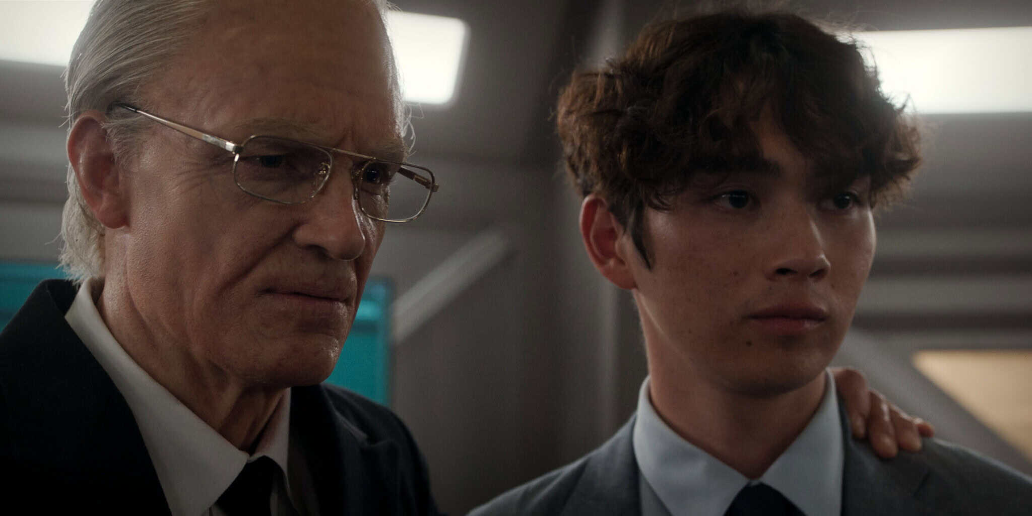 An older man with glasses and a serious expression stands beside a young man in a suit, resting his hand on the young man’s shoulder. Both look ahead with focused expressions in a well-lit indoor setting.