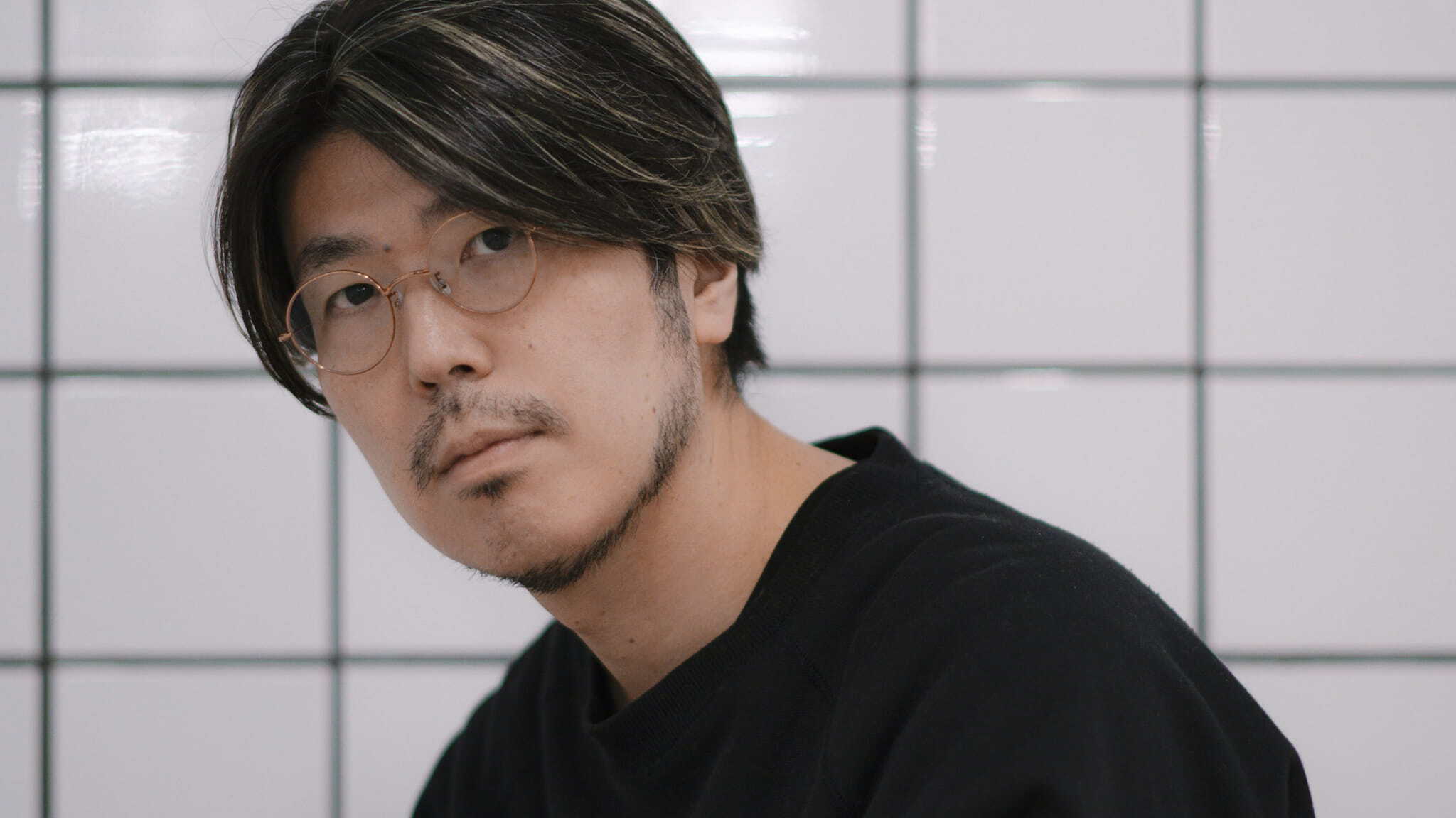 A man with glasses and medium-length dark hair, wearing a black shirt, looks at the camera in front of a white tiled wall with black grid lines.