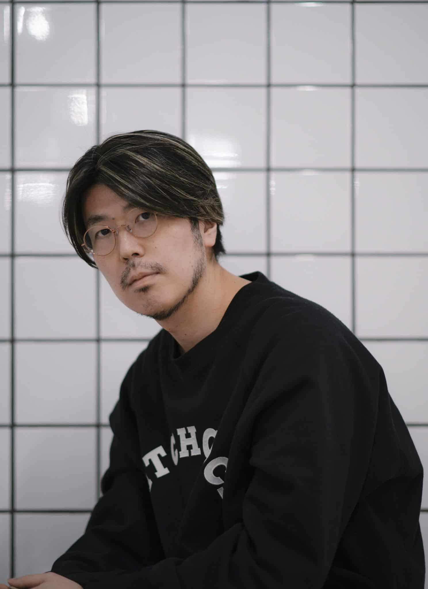 A man with glasses and medium-length dark hair, wearing a black shirt, looks at the camera in front of a white tiled wall with black grid lines.