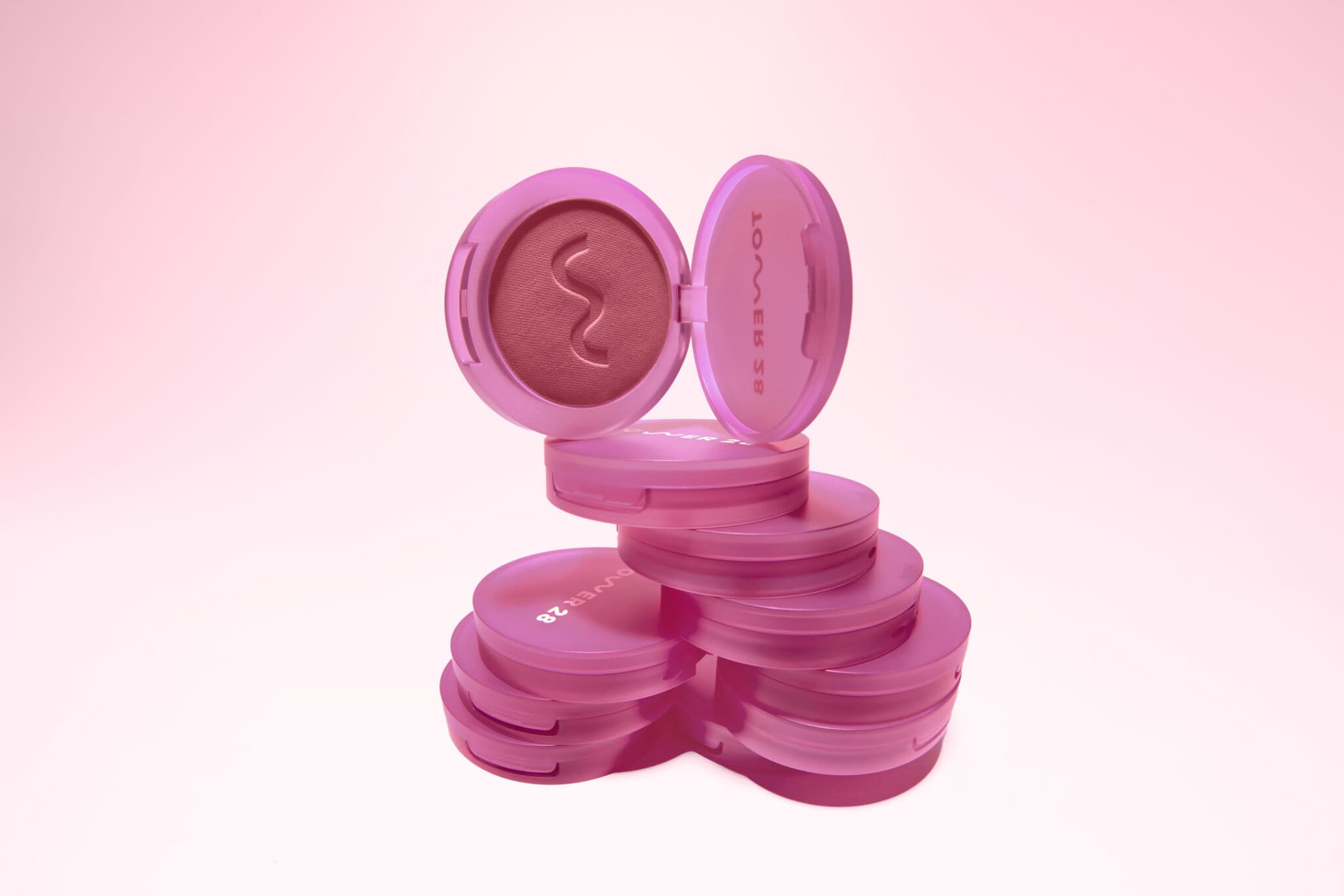A stack of pink round makeup compacts, with one compact open to reveal mauve powder and a wavy design. The background is a soft pink gradient, creating a feminine and stylish look.