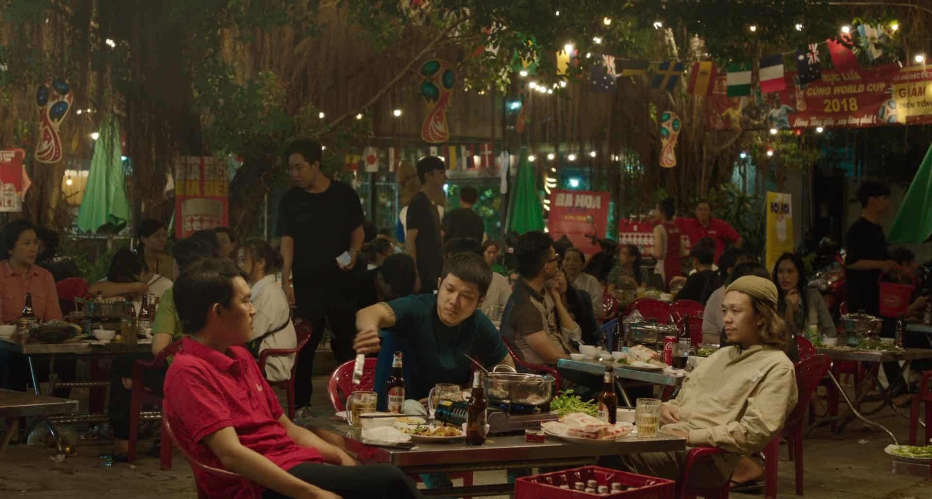 Outdoor night scene at a busy restaurant with many people seated at red tables, eating and talking. String lights and international flags hang above, creating a festive atmosphere. Food and drinks cover the tables.