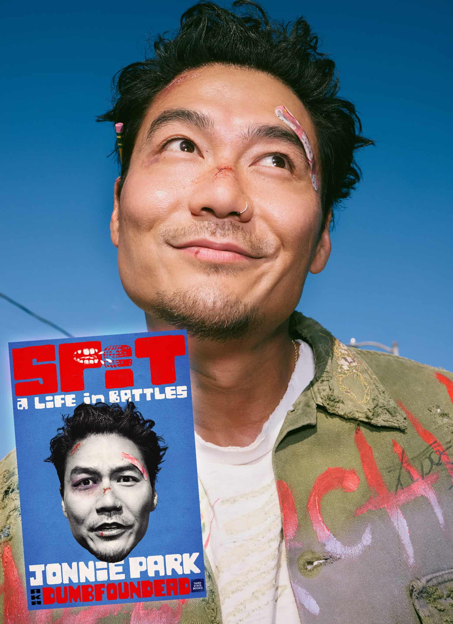A man with bandages on his face smiles against a blue background. Next to him is a poster with his face, red bold text SPIT, and the names Jonnie Park and Dumbfounded. The poster reads A Life in Battles.