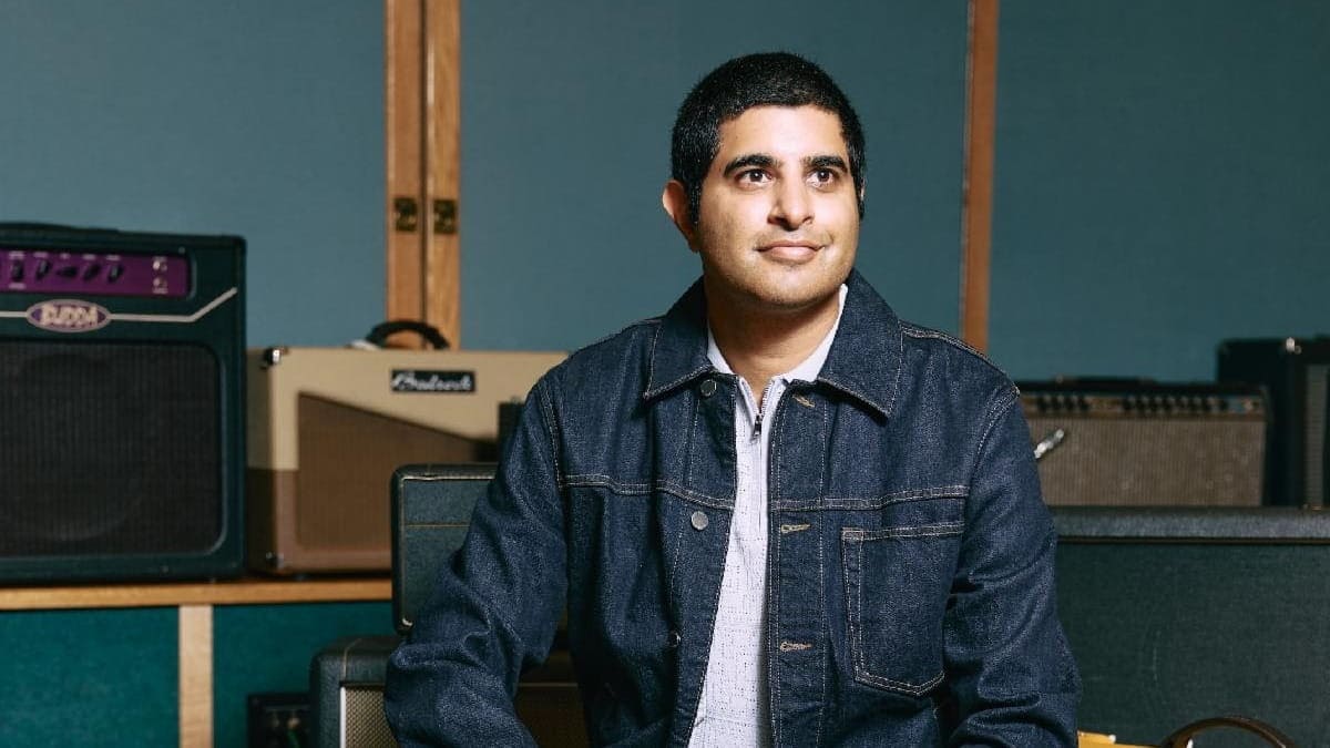 A man with short dark hair, wearing a denim jacket over a light shirt, sits in a music studio. Guitar amplifiers are visible behind him against blue and wooden walls.