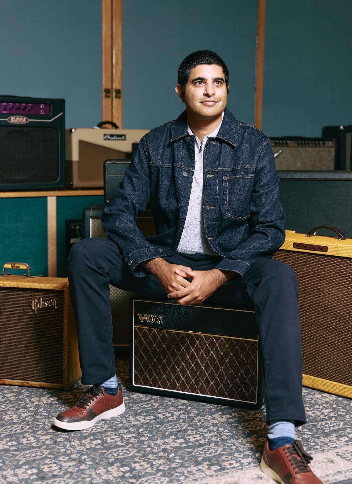 A man with short dark hair, wearing a denim jacket over a light shirt, sits in a music studio. Guitar amplifiers are visible behind him against blue and wooden walls.