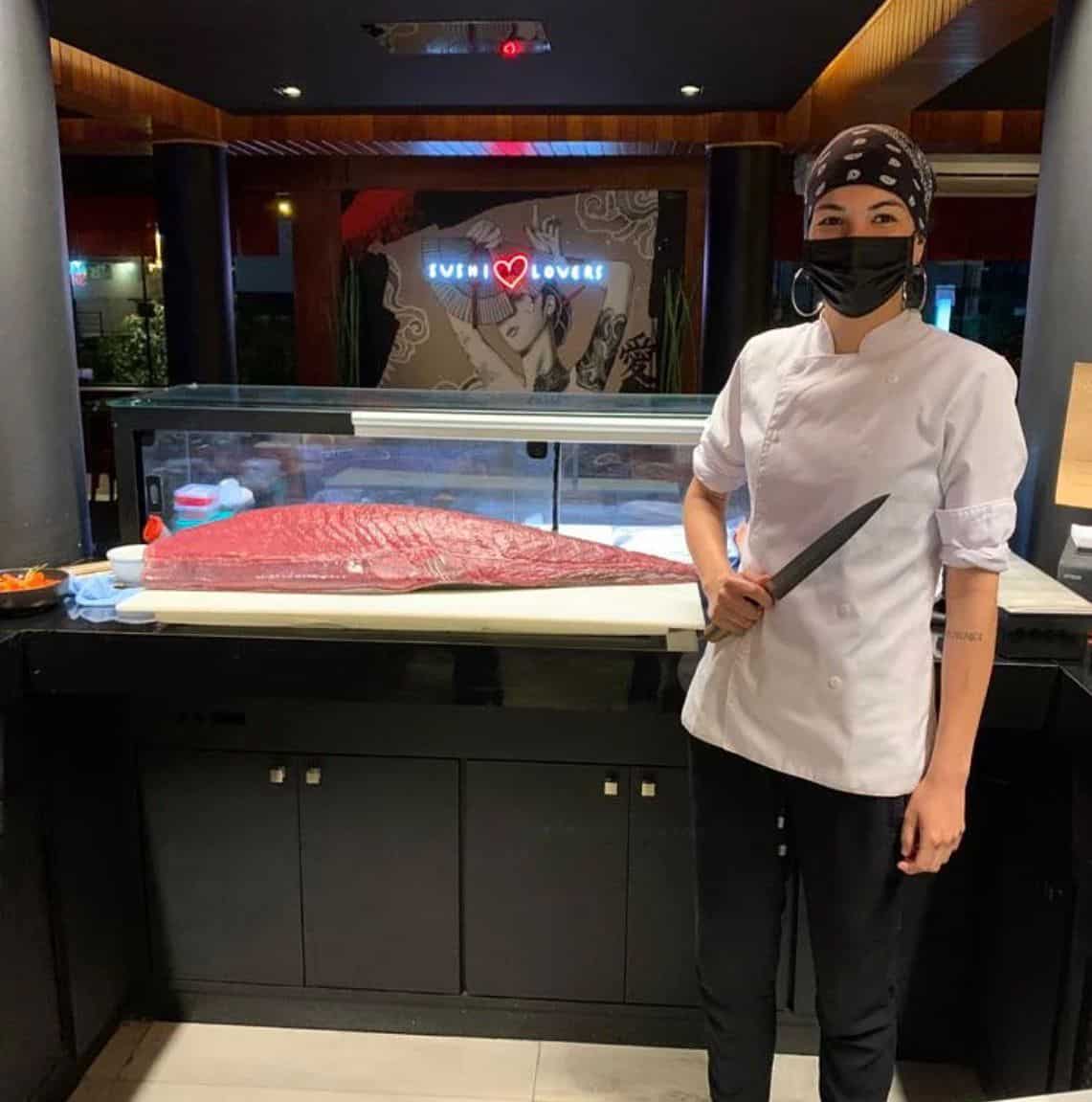 A chef wearing a white coat, black pants, headscarf, and face mask stands next to a large cut of raw fish at a sushi restaurant, holding a knife. The sign behind reads Sushi Lovers with a drawing of a woman.