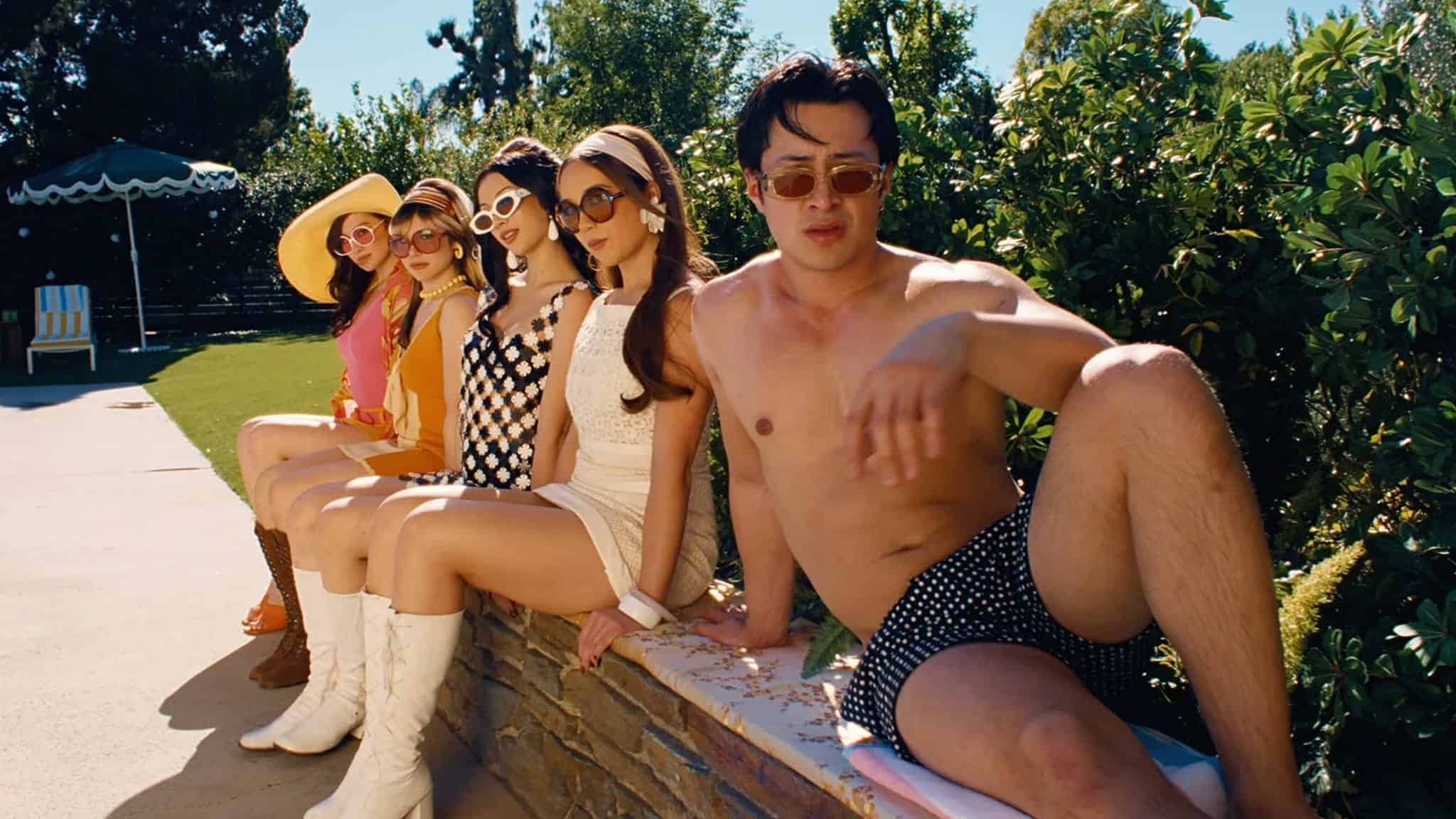 Five people in retro outfits sit by a pool; four women in colorful, vintage dresses and boots, and one shirtless man in swim trunks, all wearing sunglasses and looking relaxed in the sun.