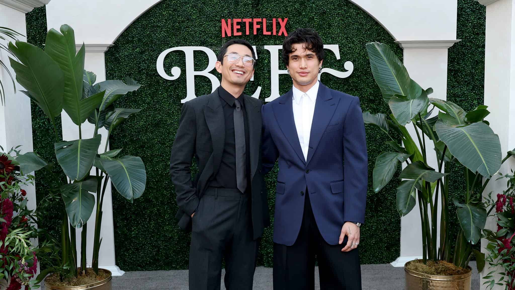 Two men dressed in suits stand and smile in front of a green hedge backdrop with “Netflix” and “BEEF” written on it, surrounded by large potted plants.