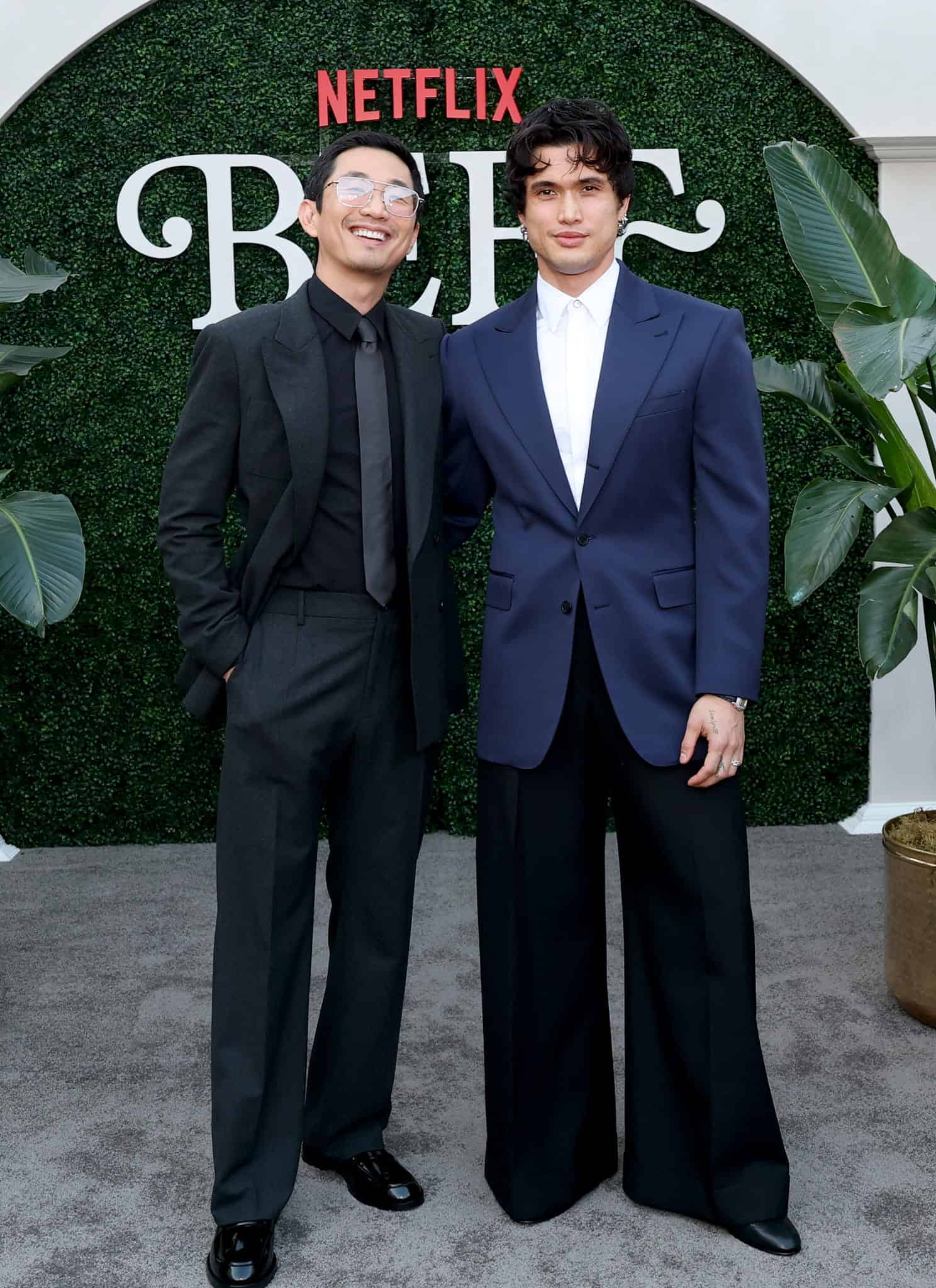 Two men dressed in suits stand and smile in front of a green hedge backdrop with “Netflix” and “BEEF” written on it, surrounded by large potted plants.