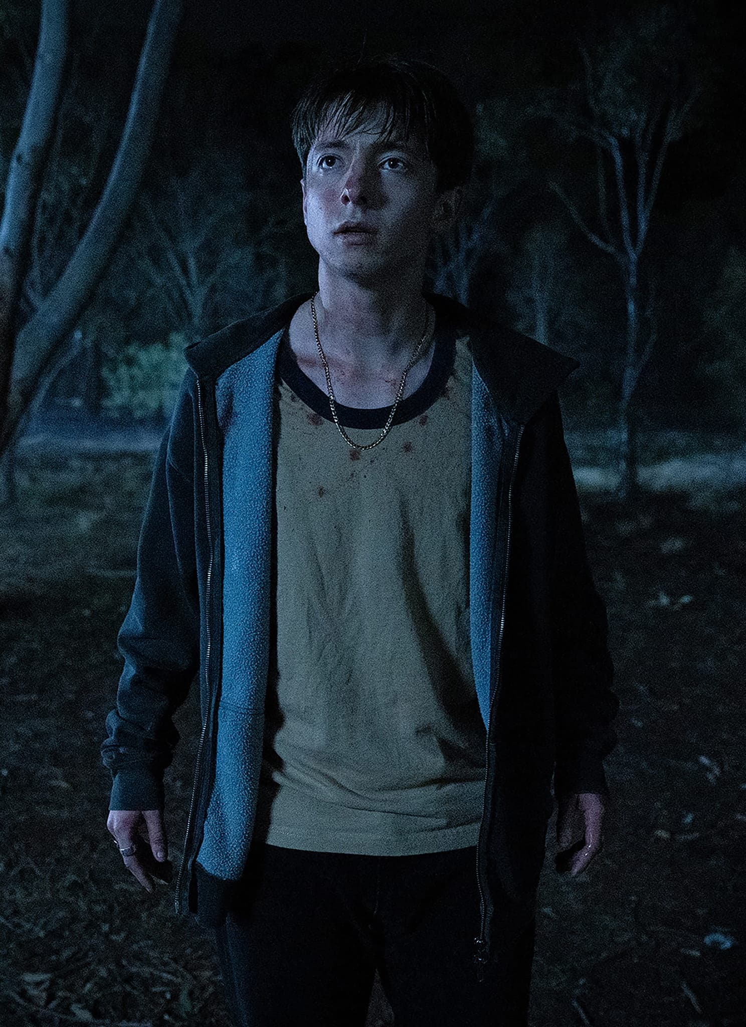 A young person stands alone in a dark, wooded area at night, looking slightly upwards with a tense expression, illuminated by dim, blue-tinged light among tall trees.
