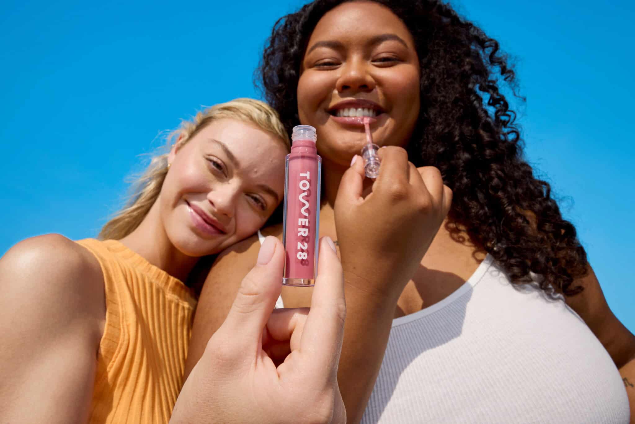 Two women smiling outdoors under a blue sky, one holding and applying pink Tower 28 lip gloss while the other poses beside her. The focus is on the lip gloss held up toward the camera.