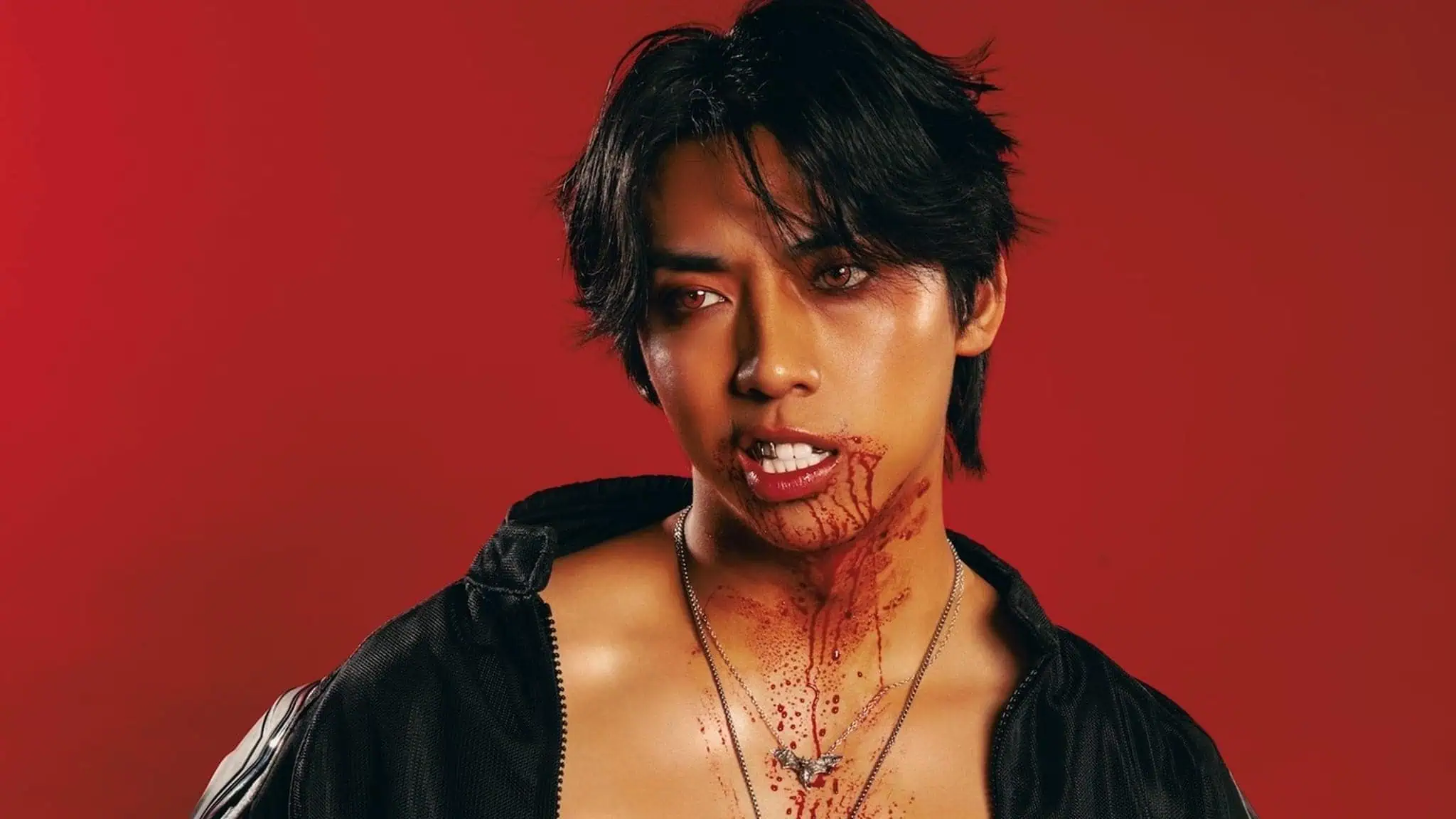 A person with dark hair and blood on their mouth, chin, and neck looks to the side against a red background, wearing a partially unzipped black top and layered necklaces.