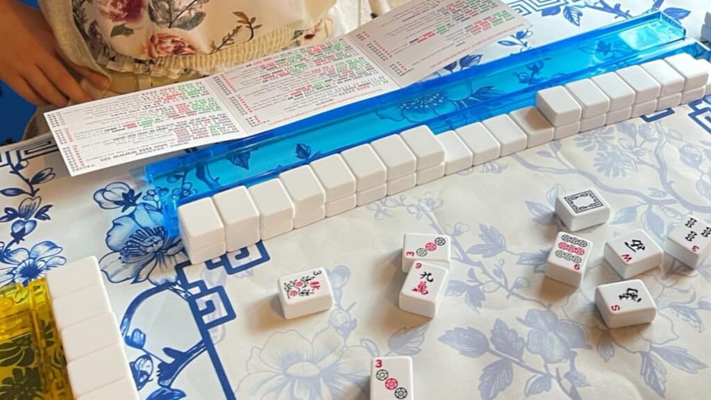 A close-up of a Mahjong game in progress on a floral-patterned table, with tiles lined up on blue and yellow racks, a scorecard, and a players hand visible near the top of the image.