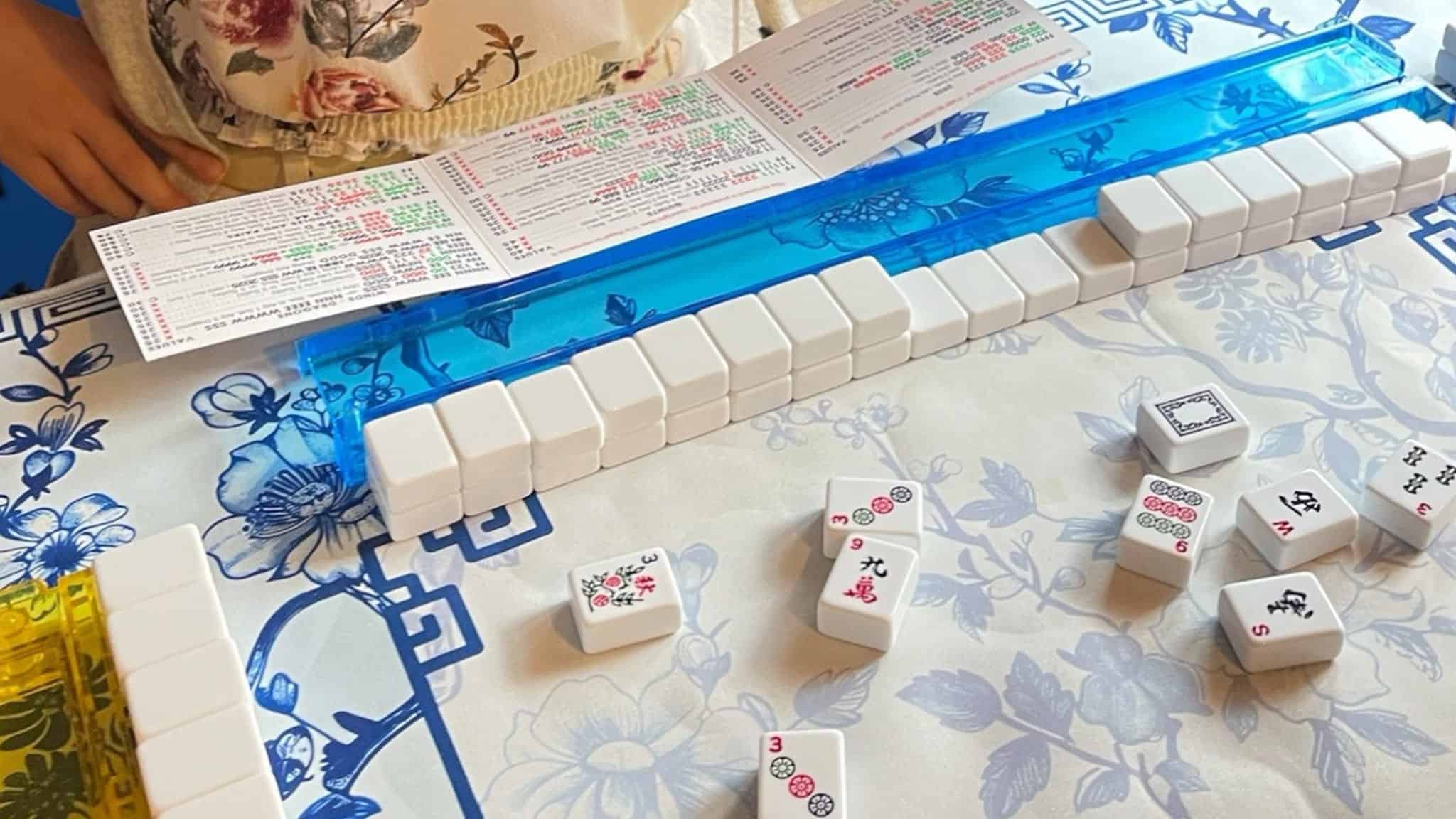 A close-up of a Mahjong game in progress on a floral-patterned table, with tiles lined up on blue and yellow racks, a scorecard, and a players hand visible near the top of the image.