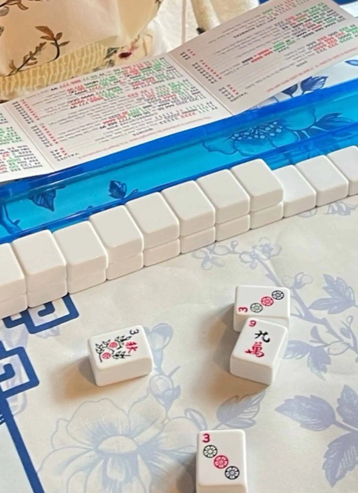 A close-up of a Mahjong game in progress on a floral-patterned table, with tiles lined up on blue and yellow racks, a scorecard, and a players hand visible near the top of the image.