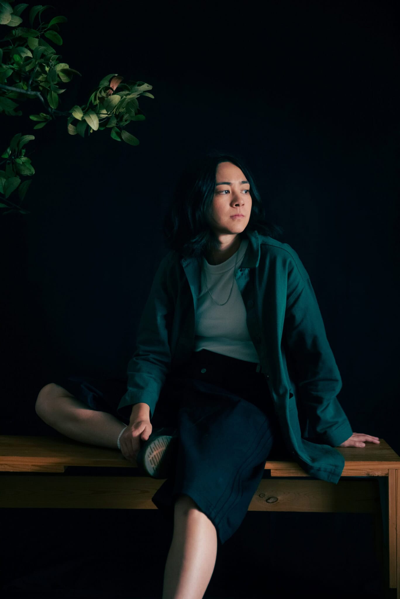 A woman with medium-length dark hair sits on a wooden table against a dark background, wearing a green jacket and black skirt. One leg is bent up on the table. Green leafy branches hang in the top left corner.