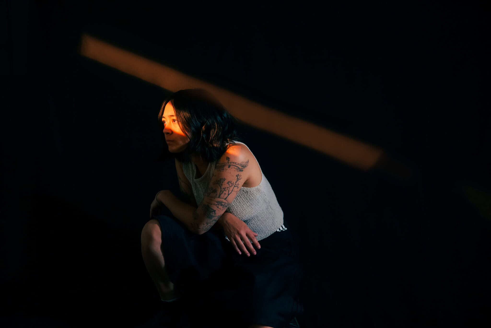 A person with dark hair and tattoos sits in a dimly lit space, illuminated by a narrow beam of light highlighting their face and upper body against a dark background.