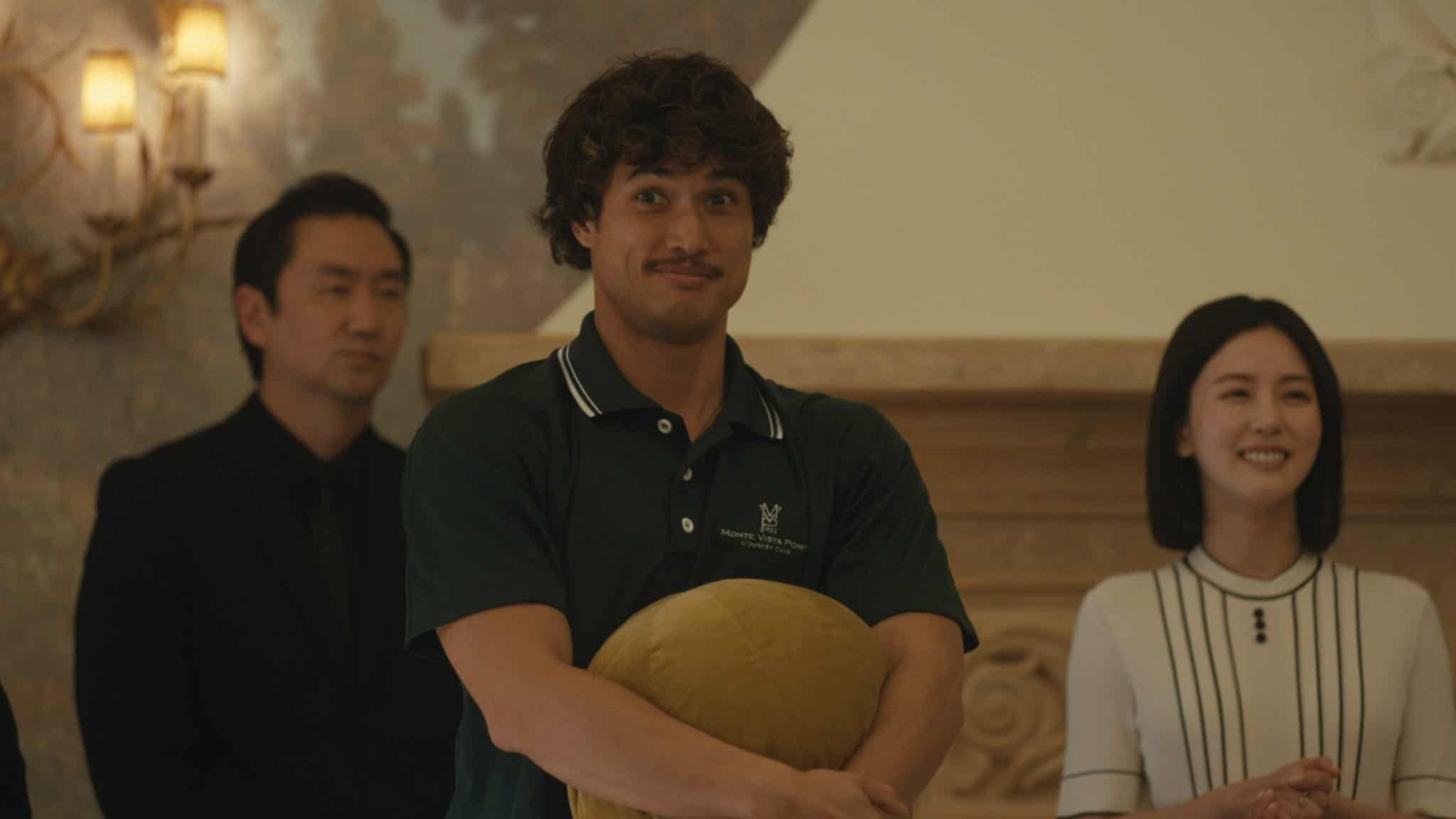 A man with curly hair and a mustache, wearing a dark green polo shirt, stands holding a yellow pillow and smiling. Two people in formal clothing stand behind him, one male and one female, both looking ahead.