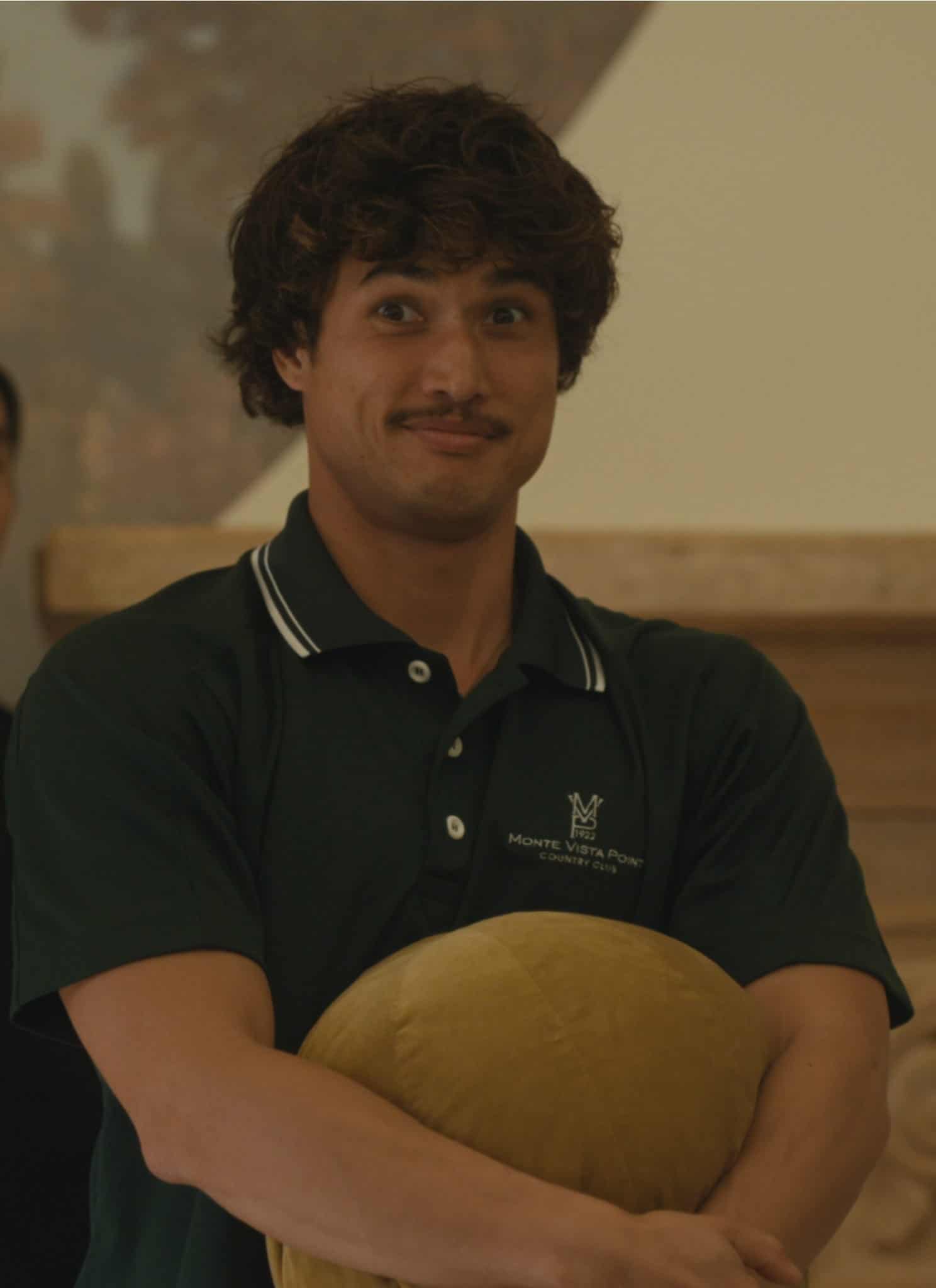 A man with curly hair and a mustache, wearing a dark green polo shirt, stands holding a yellow pillow and smiling. Two people in formal clothing stand behind him, one male and one female, both looking ahead.