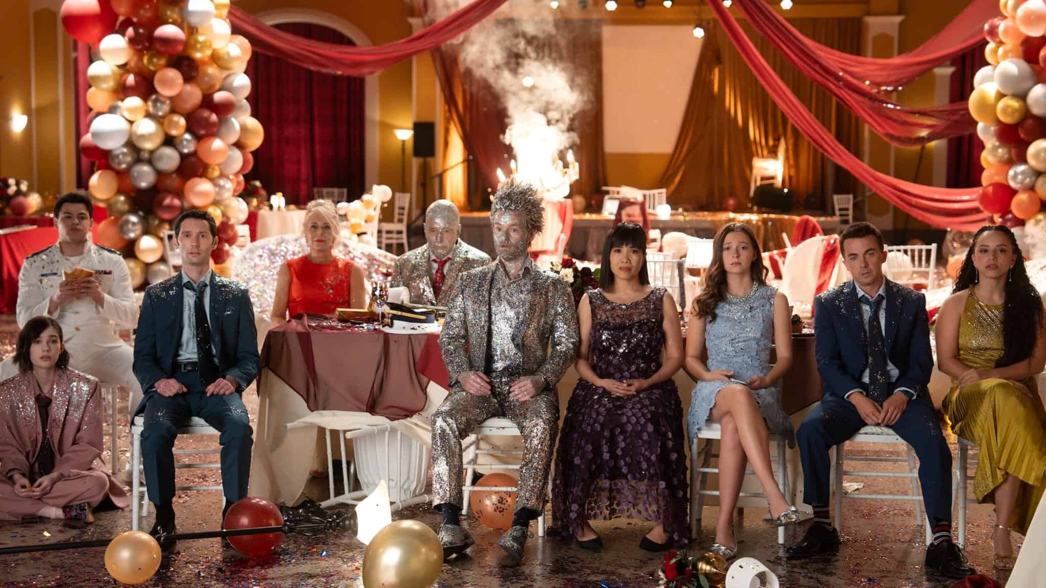 A group of people in formal attire sit in a decorated ballroom with balloons and streamers. Some look surprised or shocked, and a man in the center is covered in silver glitter. Smoke rises from a table in the background.