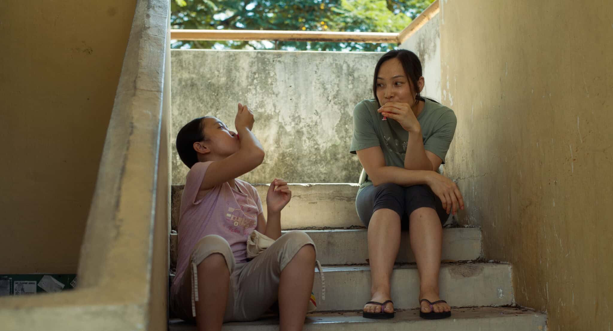 A child and an adult woman sit on outdoor concrete steps, eating snacks. The child gestures energetically while the woman looks on, smiling slightly. Sunlight and greenery are visible in the background.