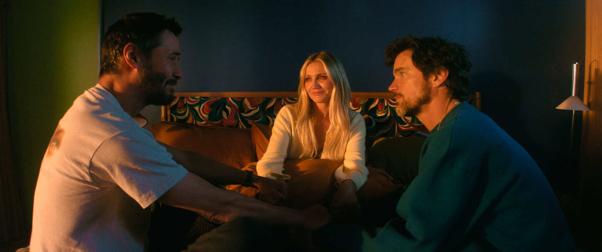 Three people sit on a bed in a warmly lit room, facing each other and smiling. The two men are on either side, and a woman sits between them, all appearing engaged in friendly conversation.