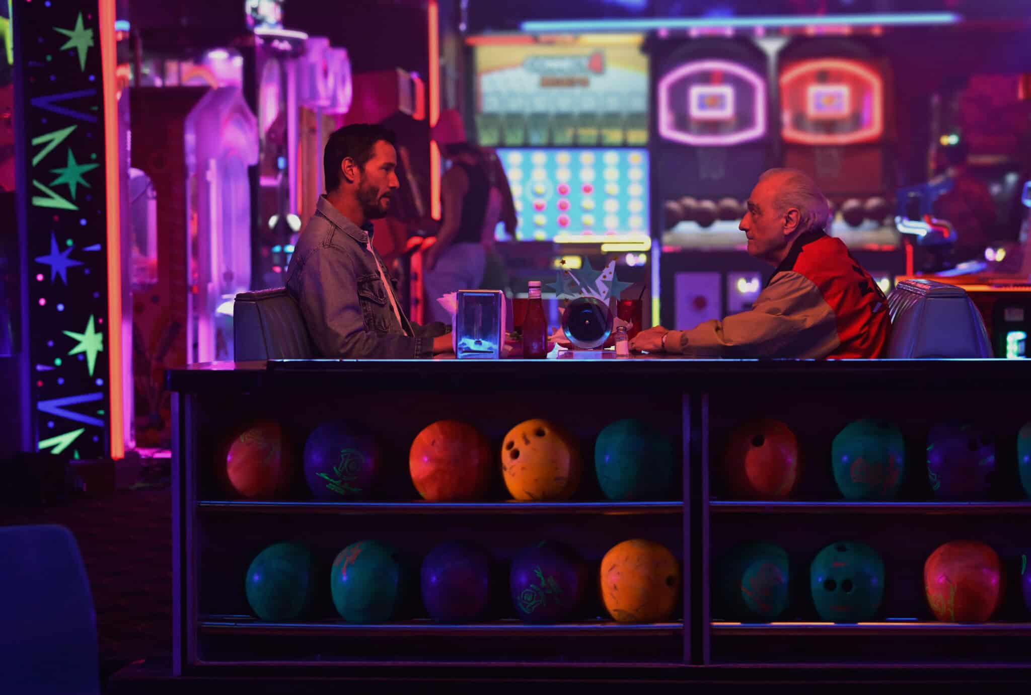 Two men sit across from each other at a table in a neon-lit bowling alley, with colorful bowling balls on a rack in the foreground and arcade games glowing in the background.