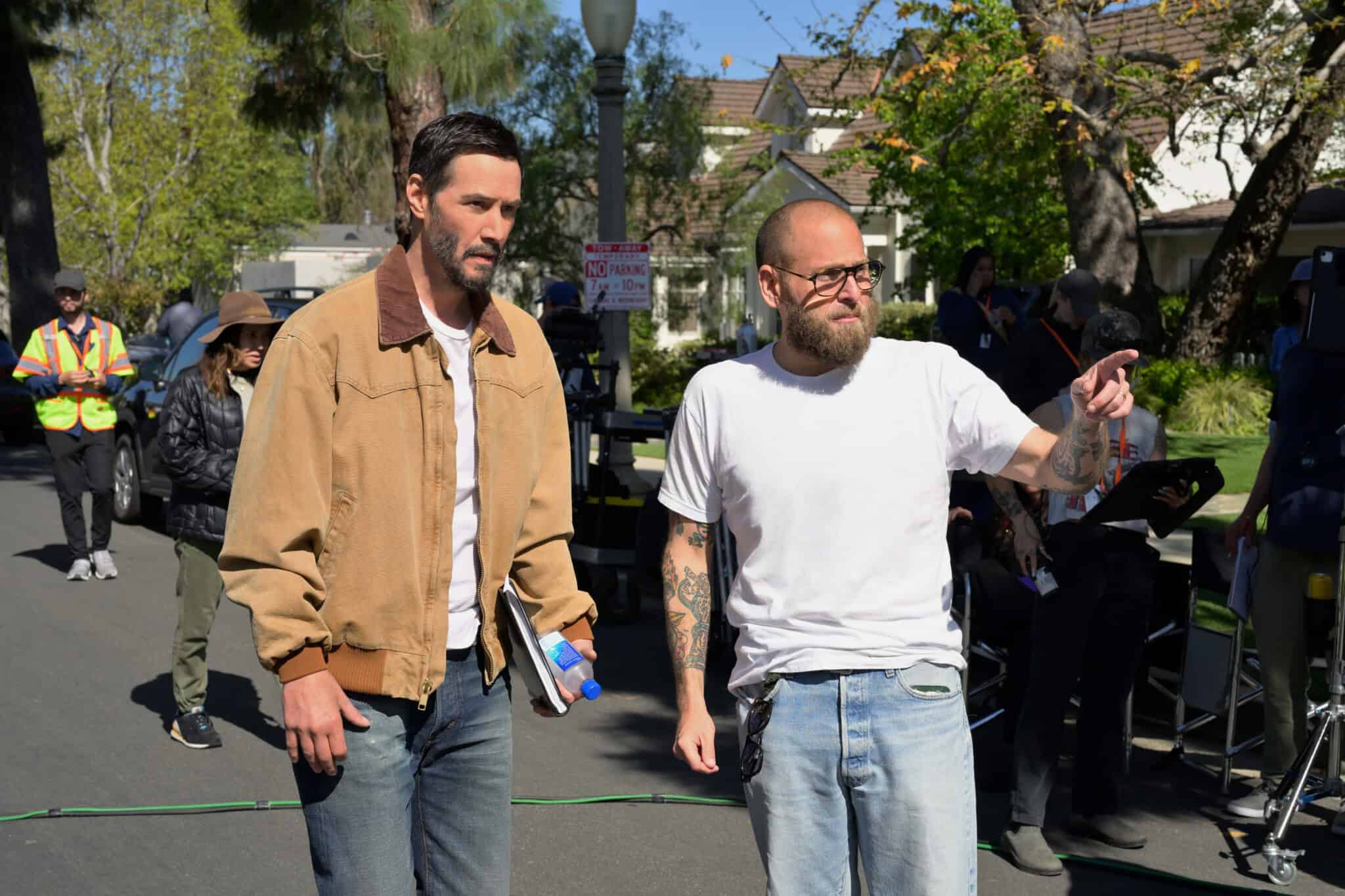 Two men stand on a suburban street film set; one wears a tan jacket and holds a notebook and water, the other in a white T-shirt gestures as if giving directions. Crew and equipment are visible in the background.