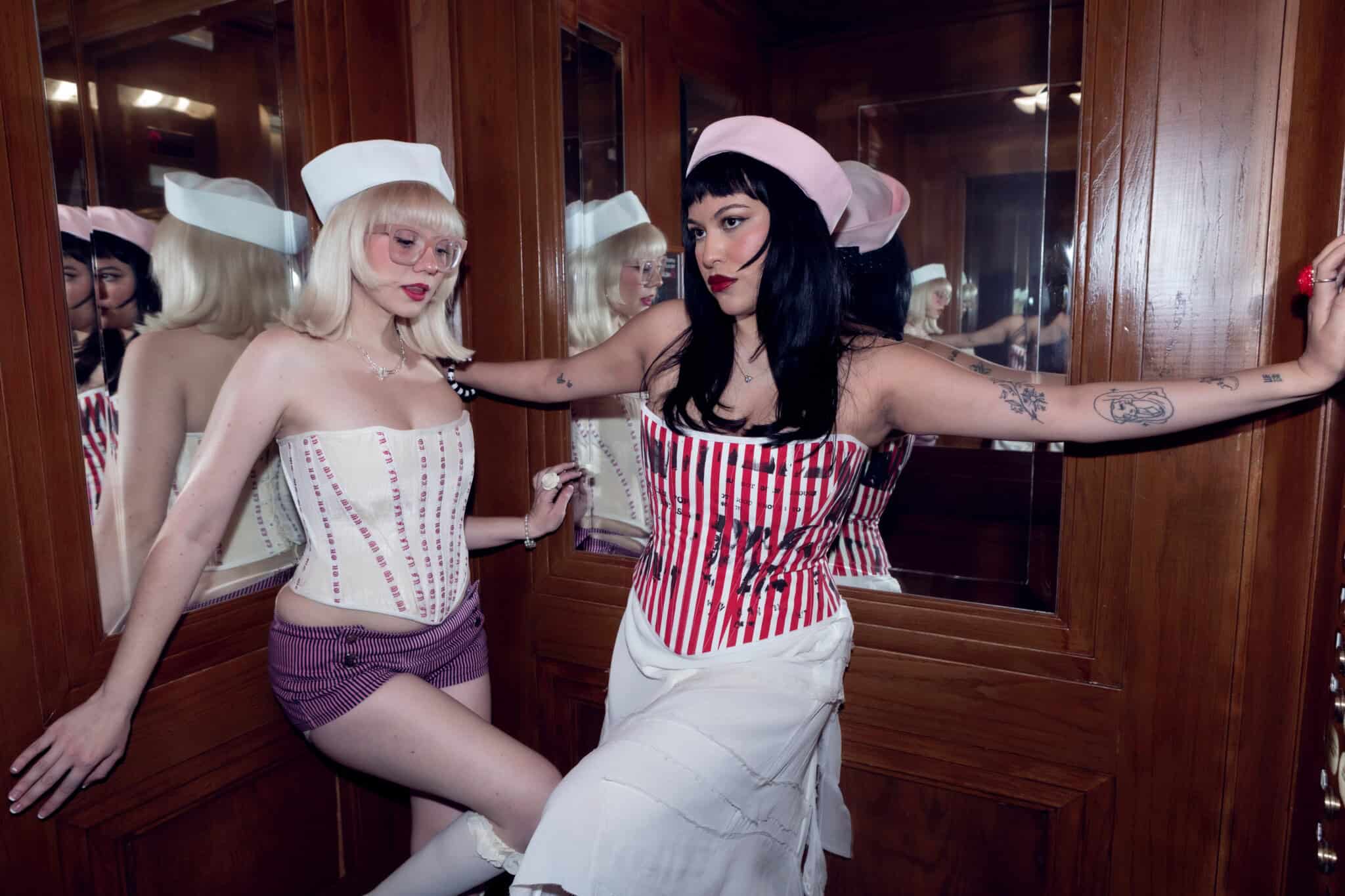 Two women pose in a wood-paneled room with mirrors. Both wear white caps and corsets, one in a white and pink outfit with glasses, and the other in a red-striped corset with white skirt and dark hair.