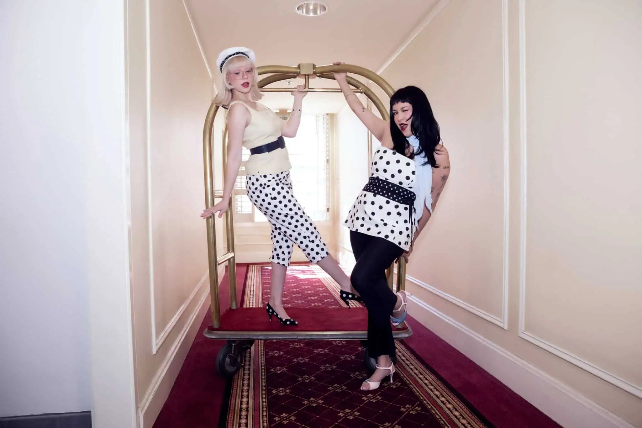 Two women pose playfully on a hotel luggage cart in a hallway. Both wear stylish, retro-inspired outfits with polka dots and hats. One stands, leaning on the cart, while the other sits on the edge, both smiling and having fun.