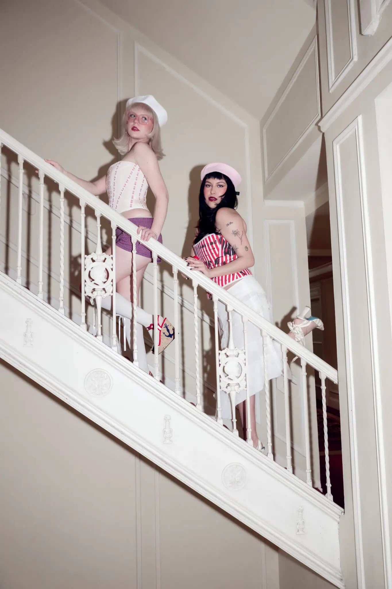 Two women in retro-inspired sailor outfits pose on a white staircase indoors. One wears a pale corset with shorts, the other a striped top with a white skirt. Both have hats and heels, giving a vintage, playful vibe.