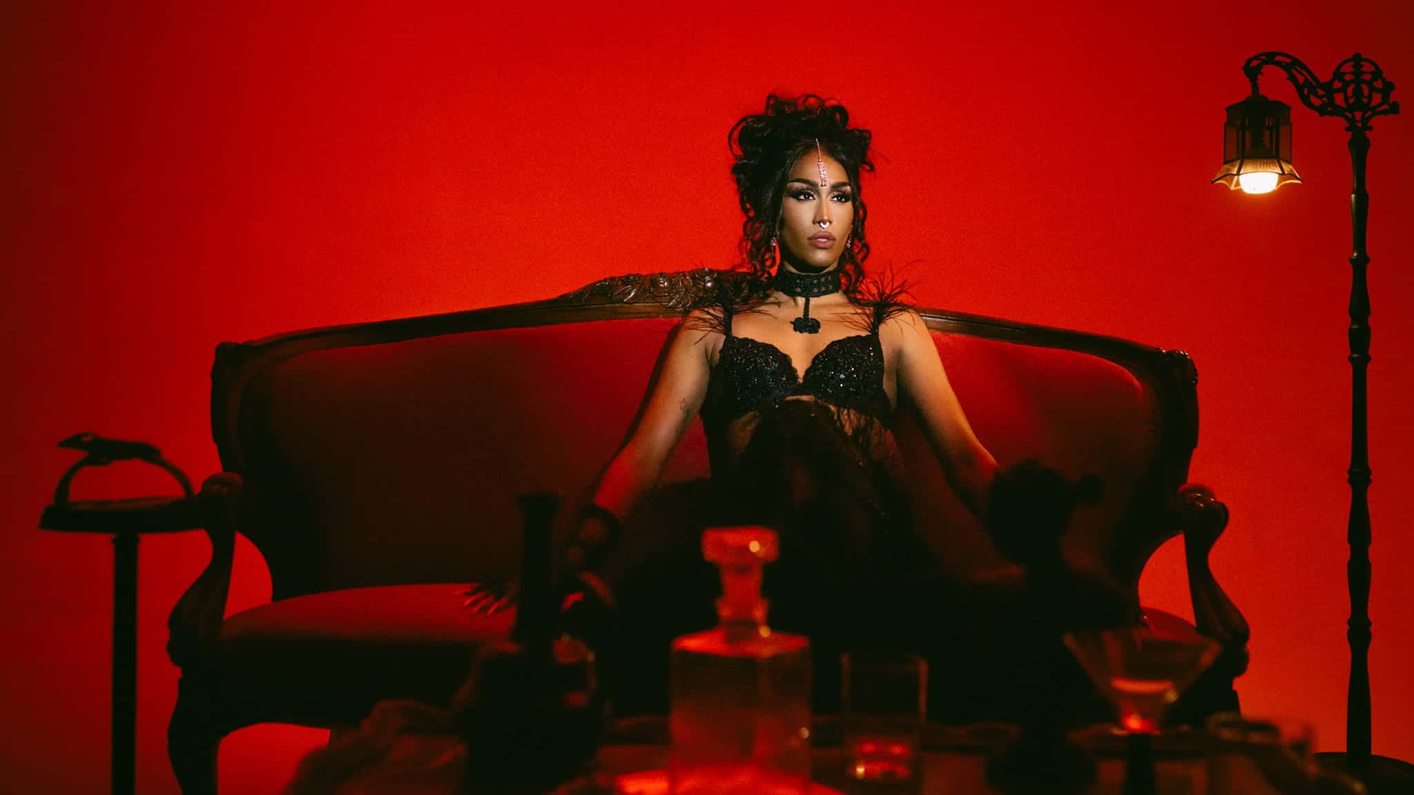 A woman in a black lace outfit sits confidently on an ornate red velvet sofa, surrounded by dim lighting and glass bottles, against a dramatic red background with a vintage lamp on the right.