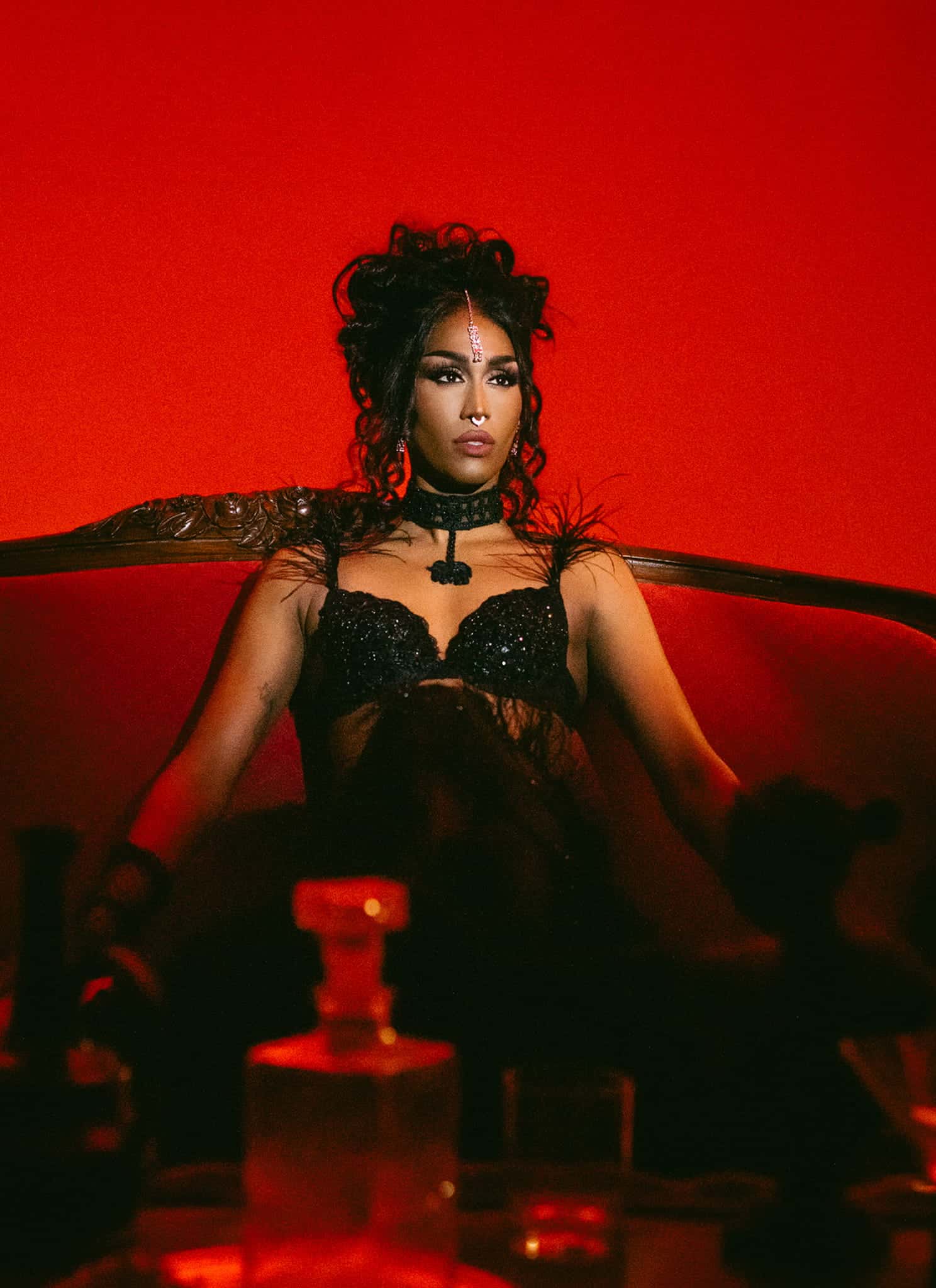 A woman in a black lace outfit sits confidently on an ornate red velvet sofa, surrounded by dim lighting and glass bottles, against a dramatic red background with a vintage lamp on the right.
