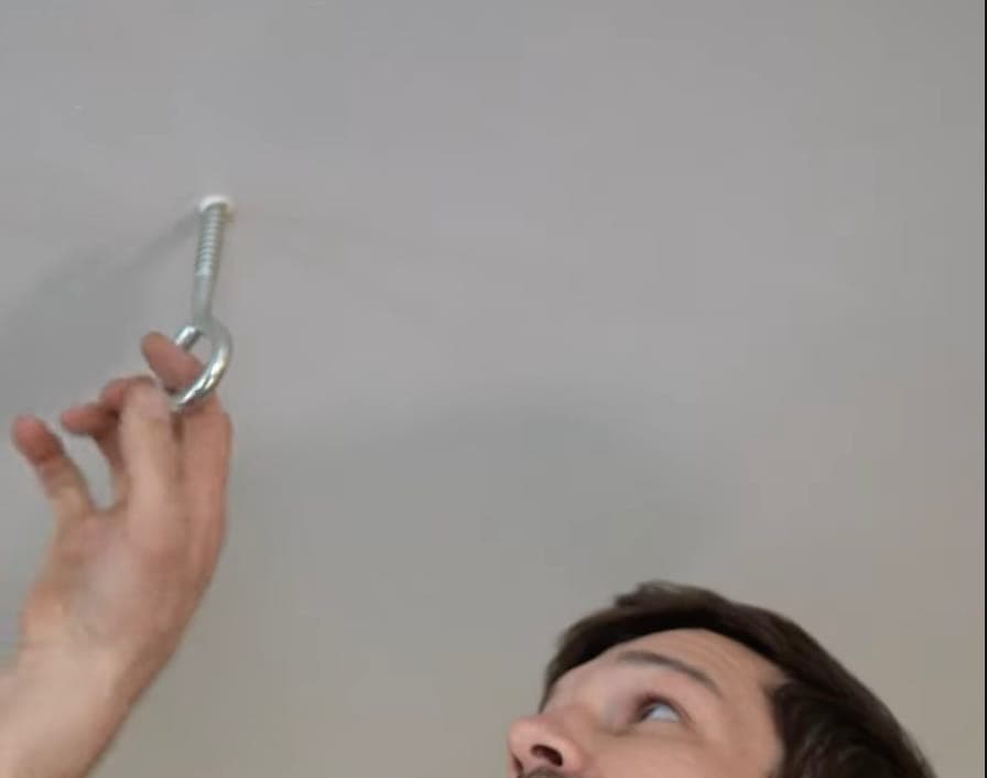 A person is looking up while screwing a metal hook into a white ceiling using their hand.