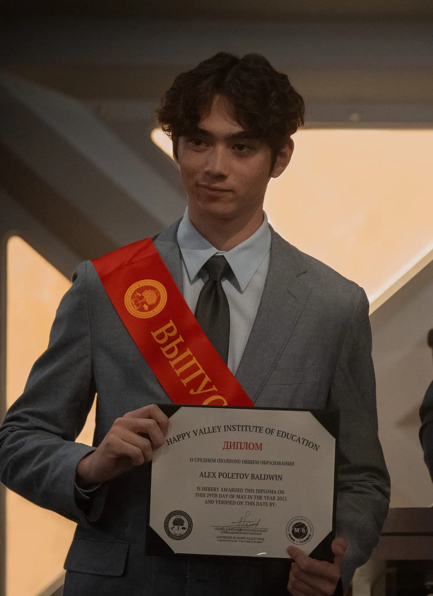 A young man in a suit holds a diploma and wears a red graduation sash, standing next to an older man in a suit and tie. They both face the camera against a softly lit indoor background.