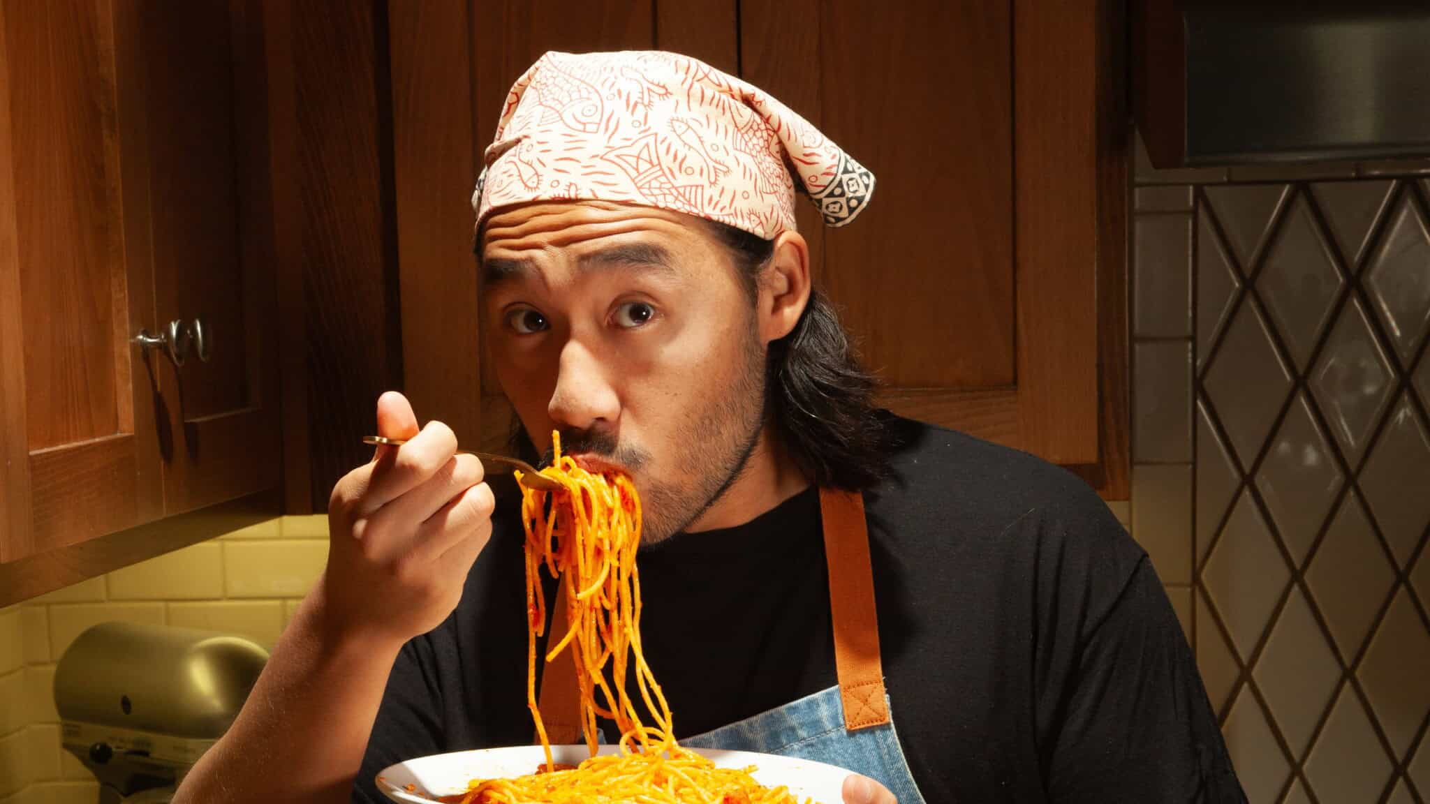A person wearing a patterned headscarf and apron is eating spaghetti from a bowl with a fork in a cozy kitchen setting with wooden cabinets.