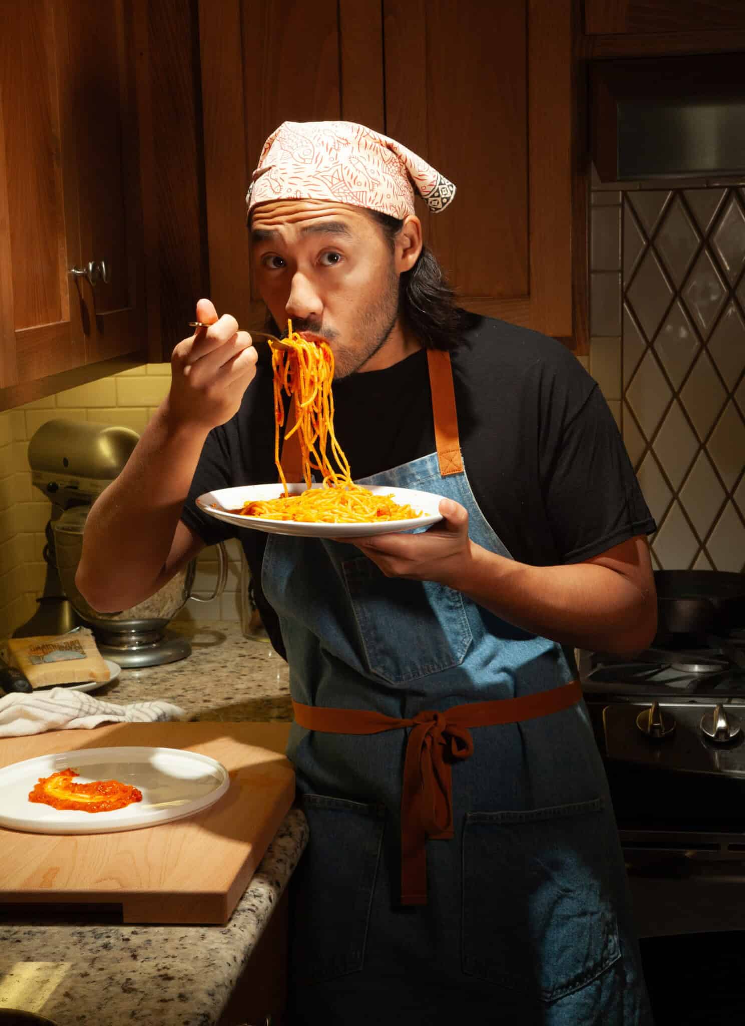 A person wearing a patterned headscarf and apron is eating spaghetti from a bowl with a fork in a cozy kitchen setting with wooden cabinets.