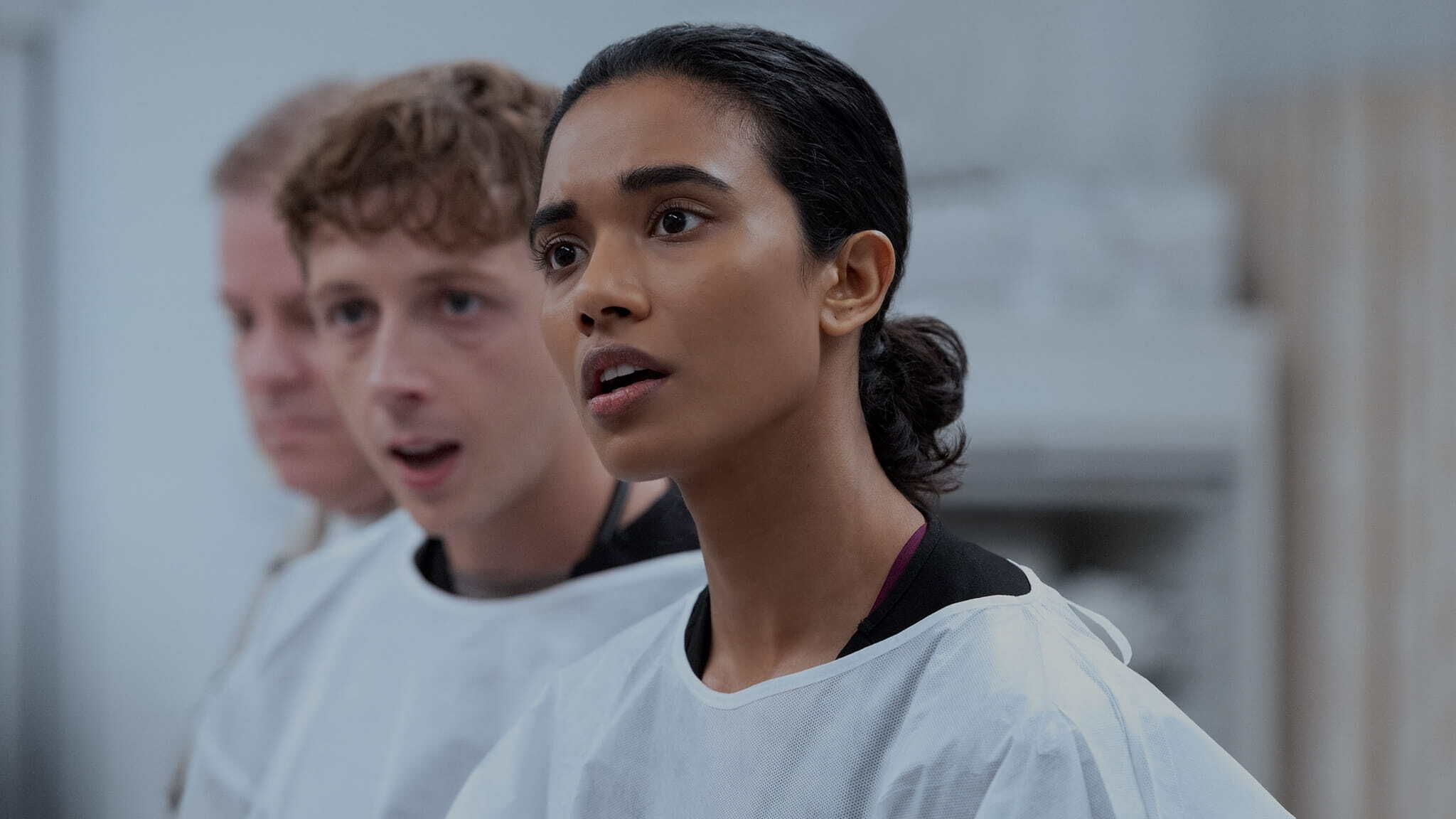 Three people wearing white lab coats stand indoors, looking ahead with focused and attentive expressions; the woman in the foreground appears to be listening intently.