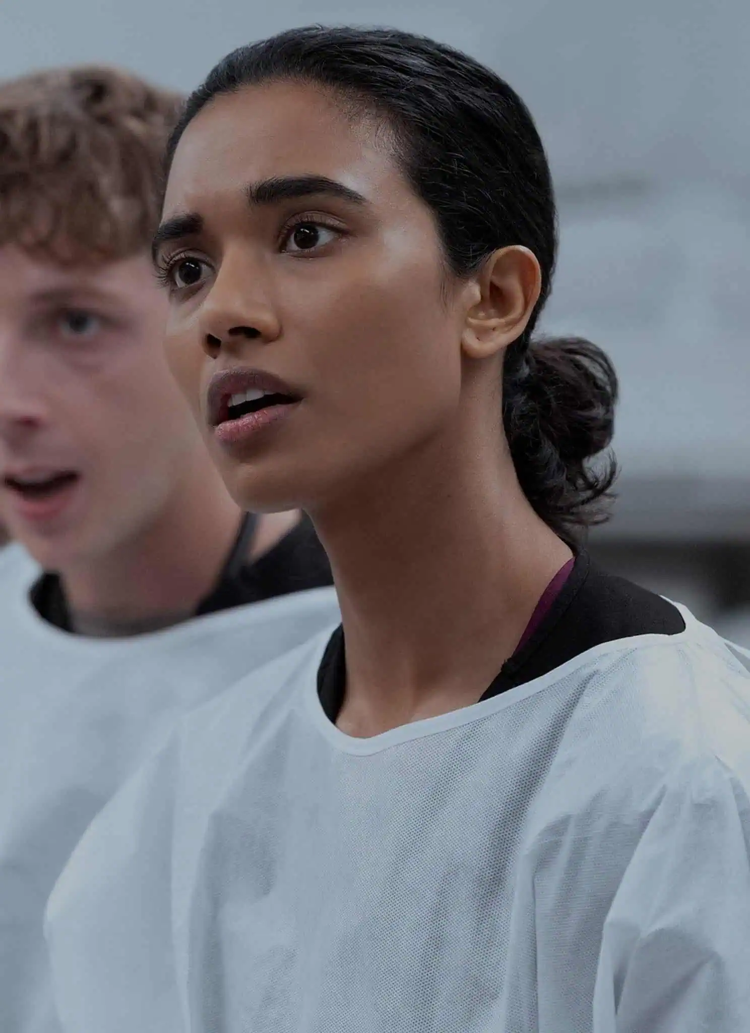 Three people wearing white lab coats stand indoors, looking ahead with focused and attentive expressions; the woman in the foreground appears to be listening intently.