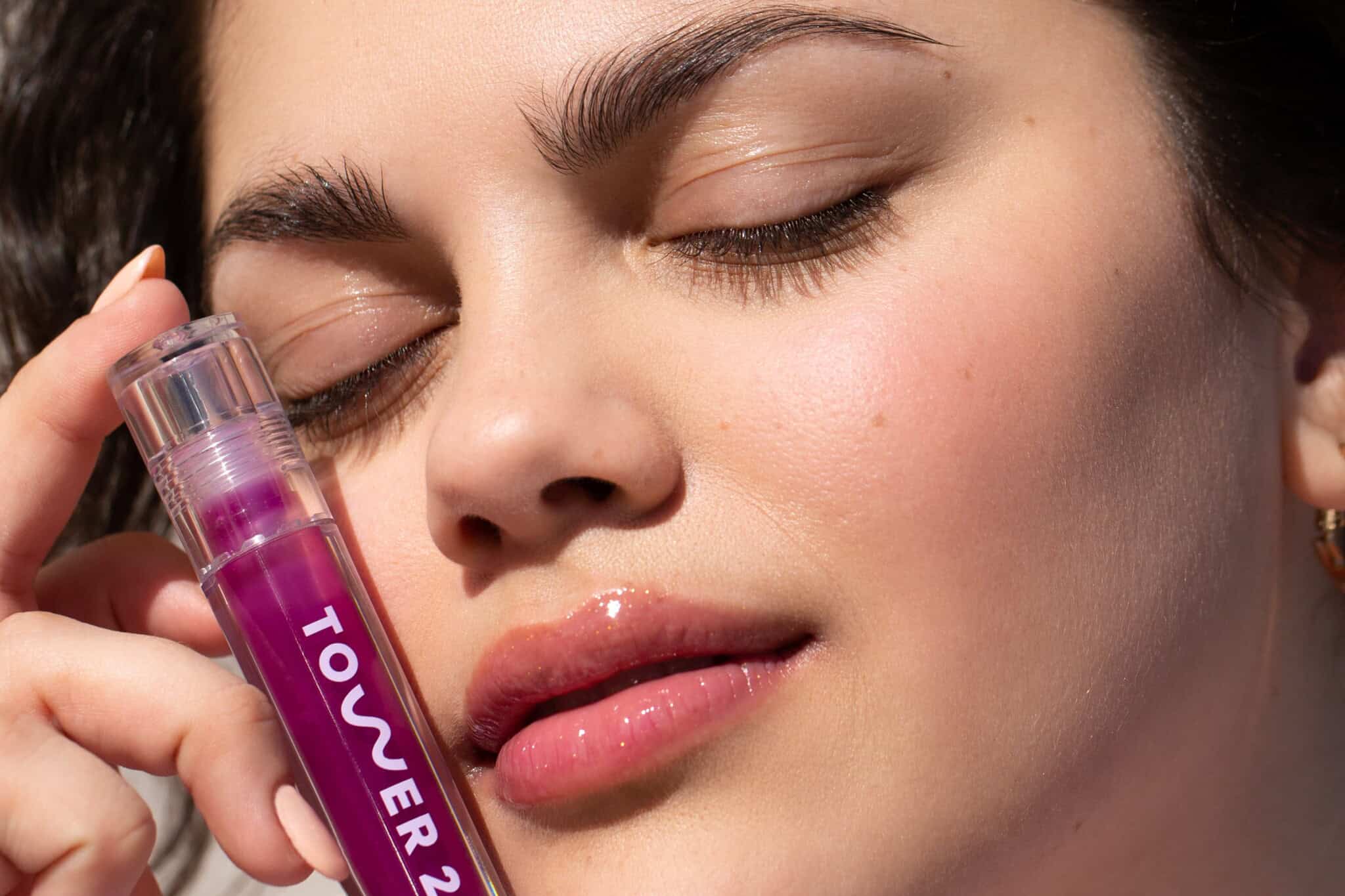 A woman with closed eyes holds a purple TOWER 28 lip gloss close to her face. Her skin looks smooth and glowing, and her lips have a glossy shine.