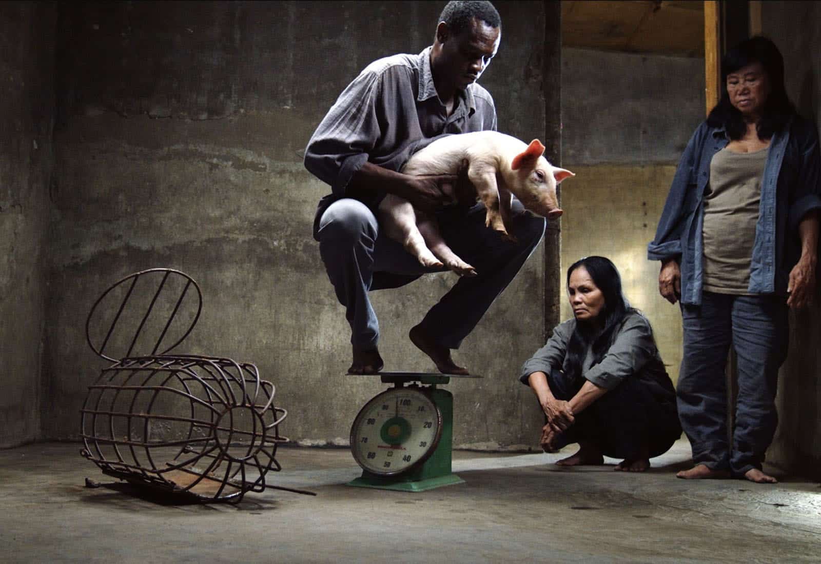 A man crouches on a scale while holding a piglet, as two women look on in a bare concrete room. An overturned metal cage lies nearby on the floor.