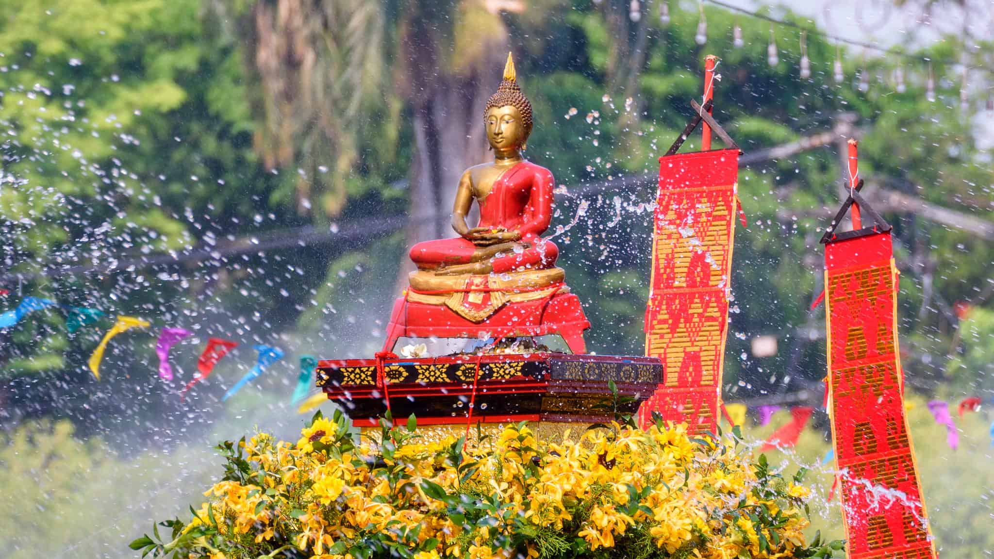 A golden Buddha statue draped in red cloth sits on a decorated pedestal surrounded by yellow flowers, with water splashing in the air and colorful flags in the background.