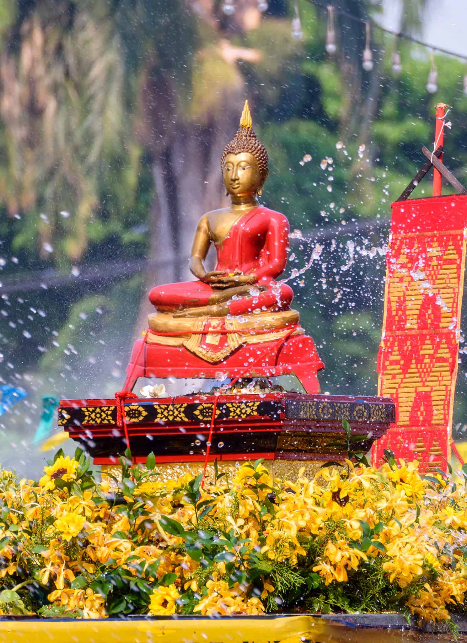 A golden Buddha statue draped in red cloth sits on a decorated pedestal surrounded by yellow flowers, with water splashing in the air and colorful flags in the background.
