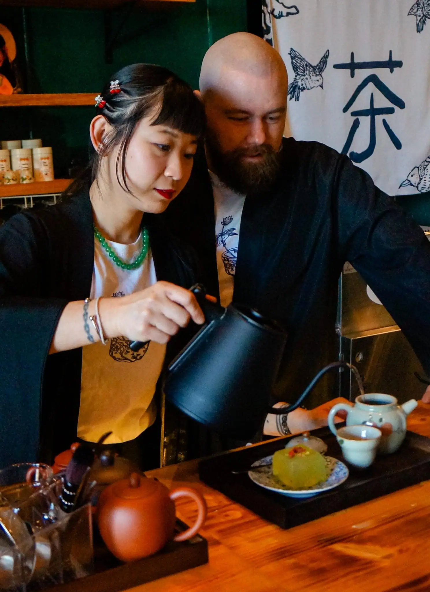 A woman and a man in traditional clothing prepare tea together at a wooden counter in a cozy teahouse, surrounded by teapots, cups, and a dessert. Japanese decor and tea utensils fill the background.