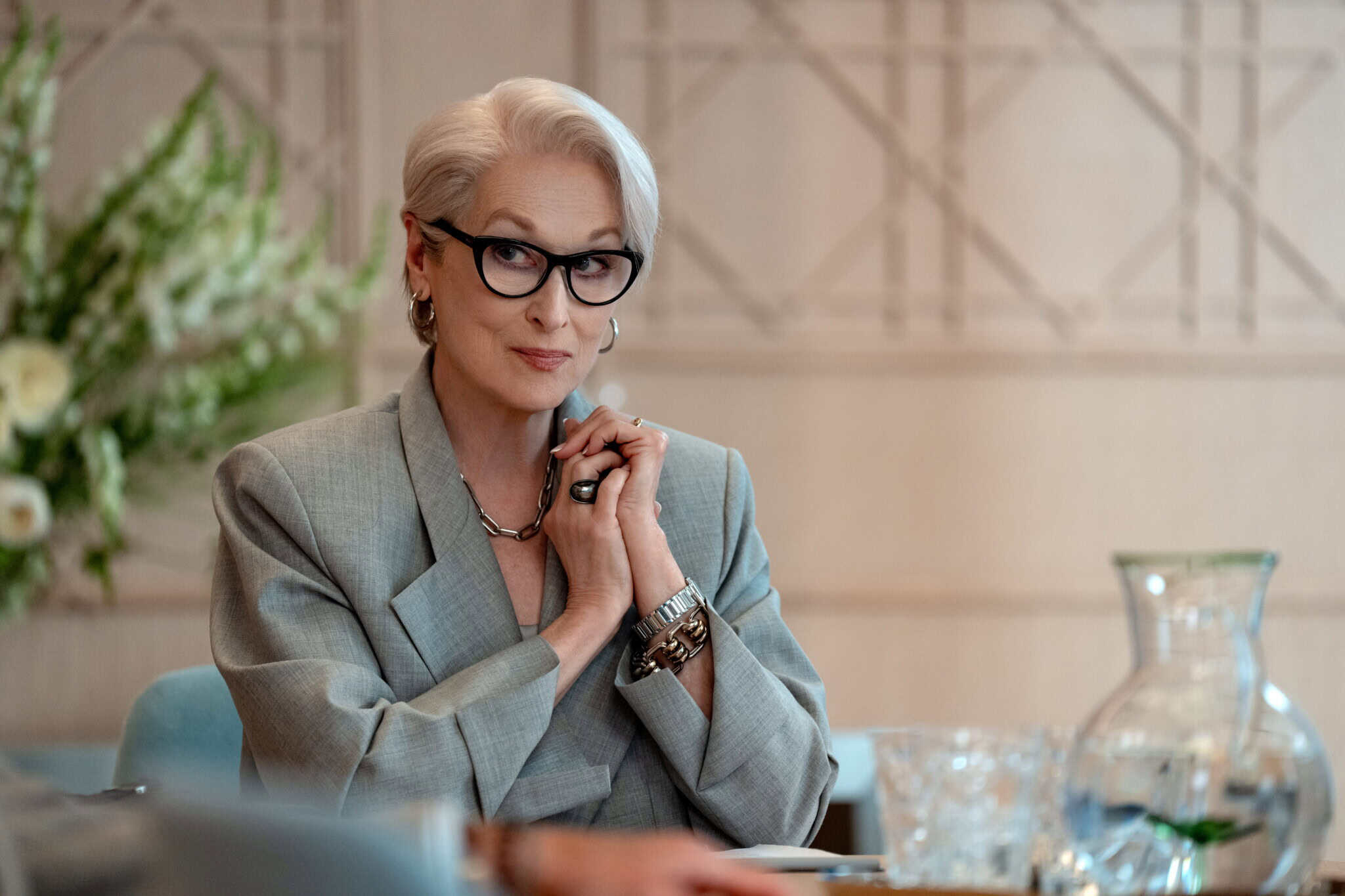 A confident woman with short white hair and glasses, wearing a gray blazer and jewelry, sits at a table with a vase and glassware, looking thoughtfully forward.