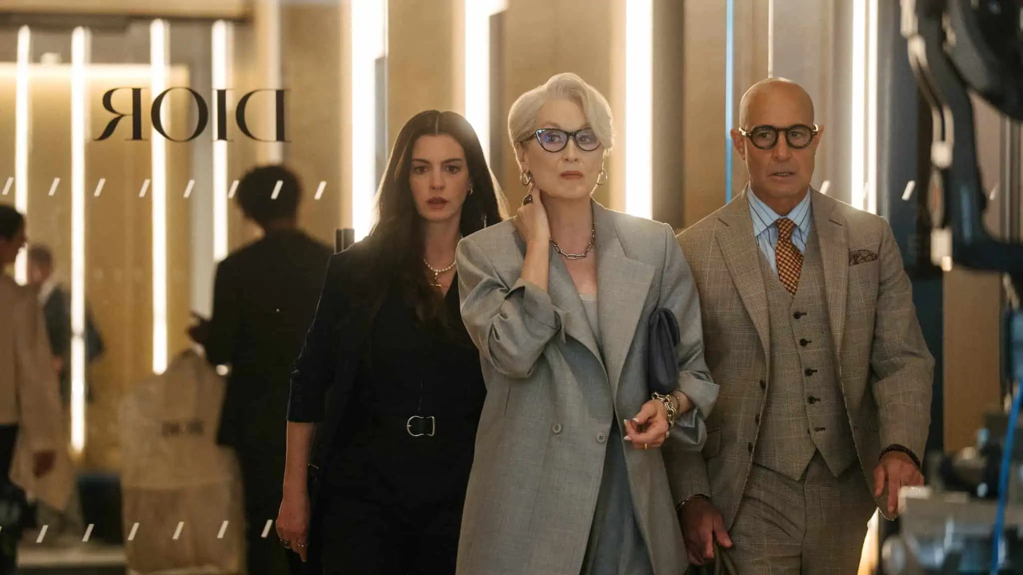 Three people, two women and one man, walk confidently through a modern, stylish hallway. The woman in the center wears a gray suit, the woman on the left wears black, and the man on the right wears a tan suit. The word “DIOR” is seen on glass.