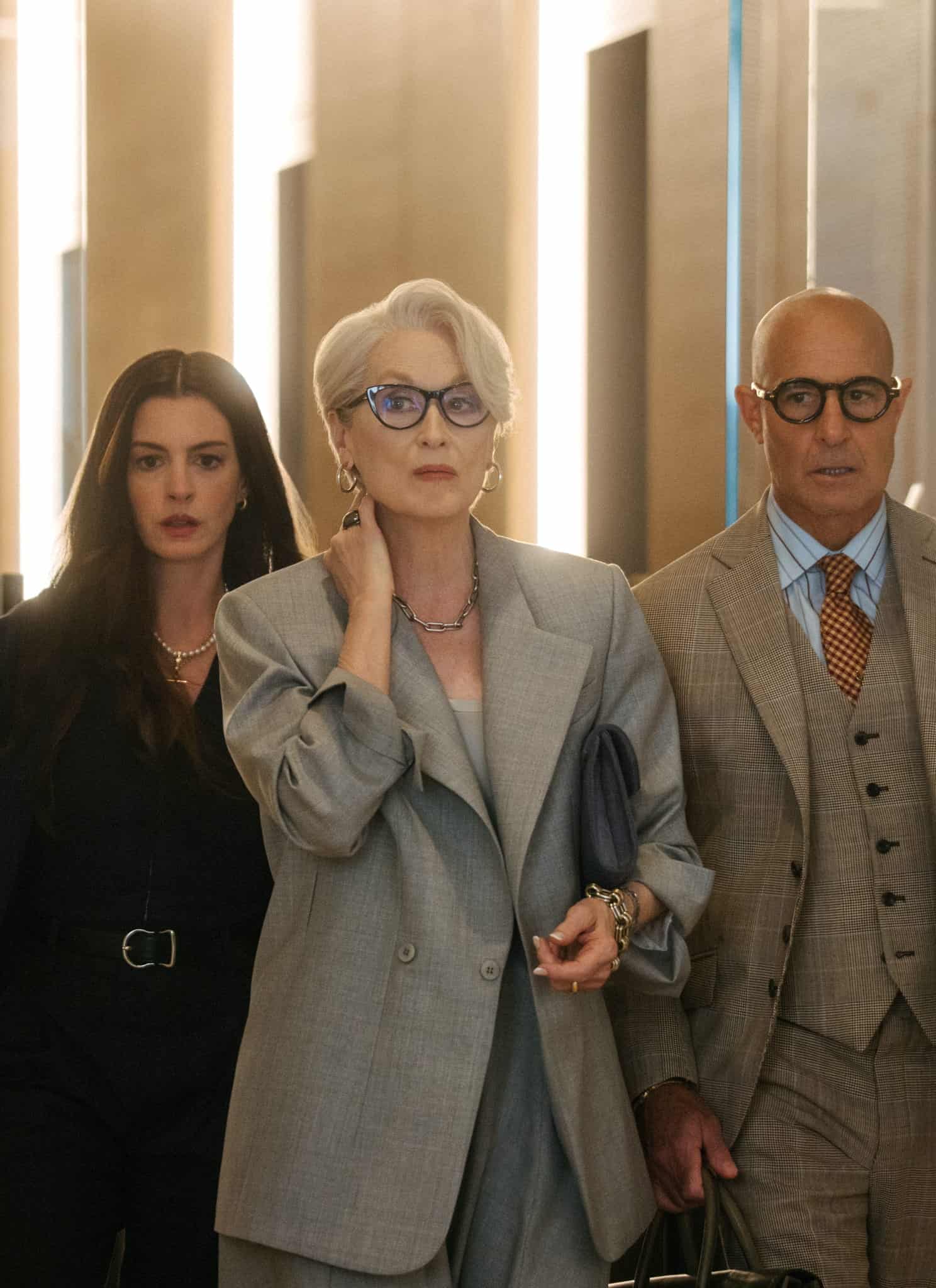 Three people, two women and one man, walk confidently through a modern, stylish hallway. The woman in the center wears a gray suit, the woman on the left wears black, and the man on the right wears a tan suit. The word “DIOR” is seen on glass.