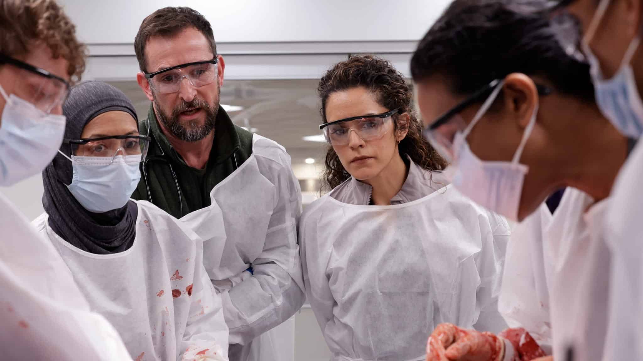A group of people wearing protective gowns, gloves, face masks, and safety goggles closely observe a procedure or demonstration, with some blood stains visible on their gowns.