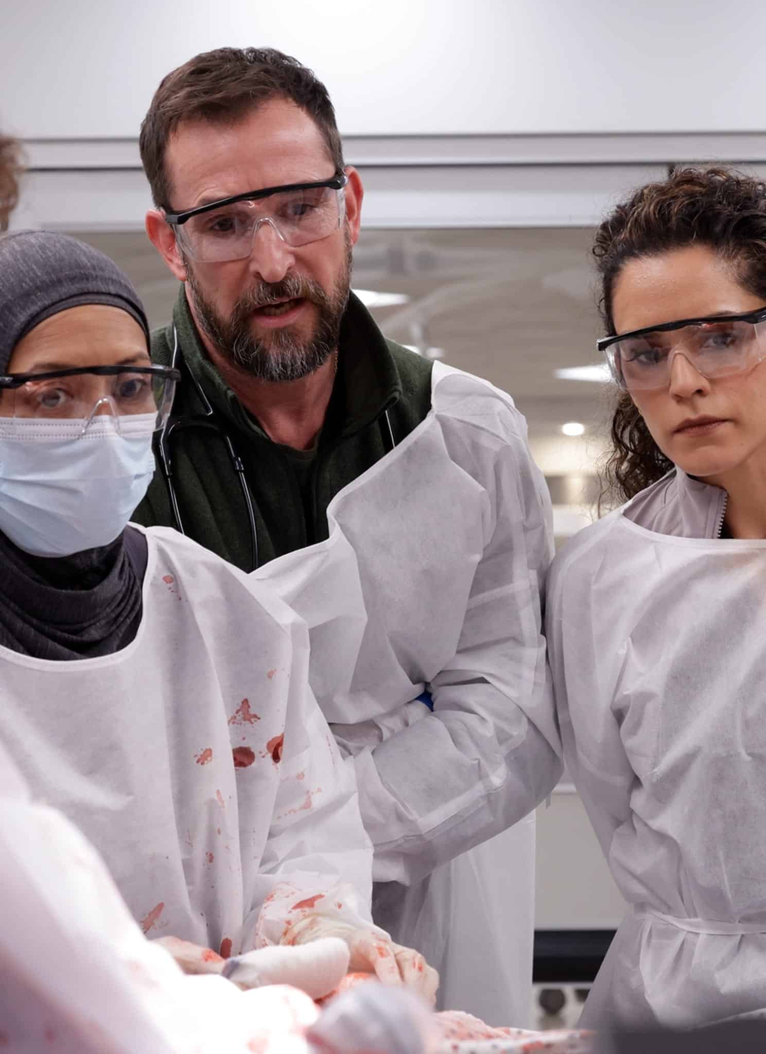 A group of people wearing protective gowns, gloves, face masks, and safety goggles closely observe a procedure or demonstration, with some blood stains visible on their gowns.