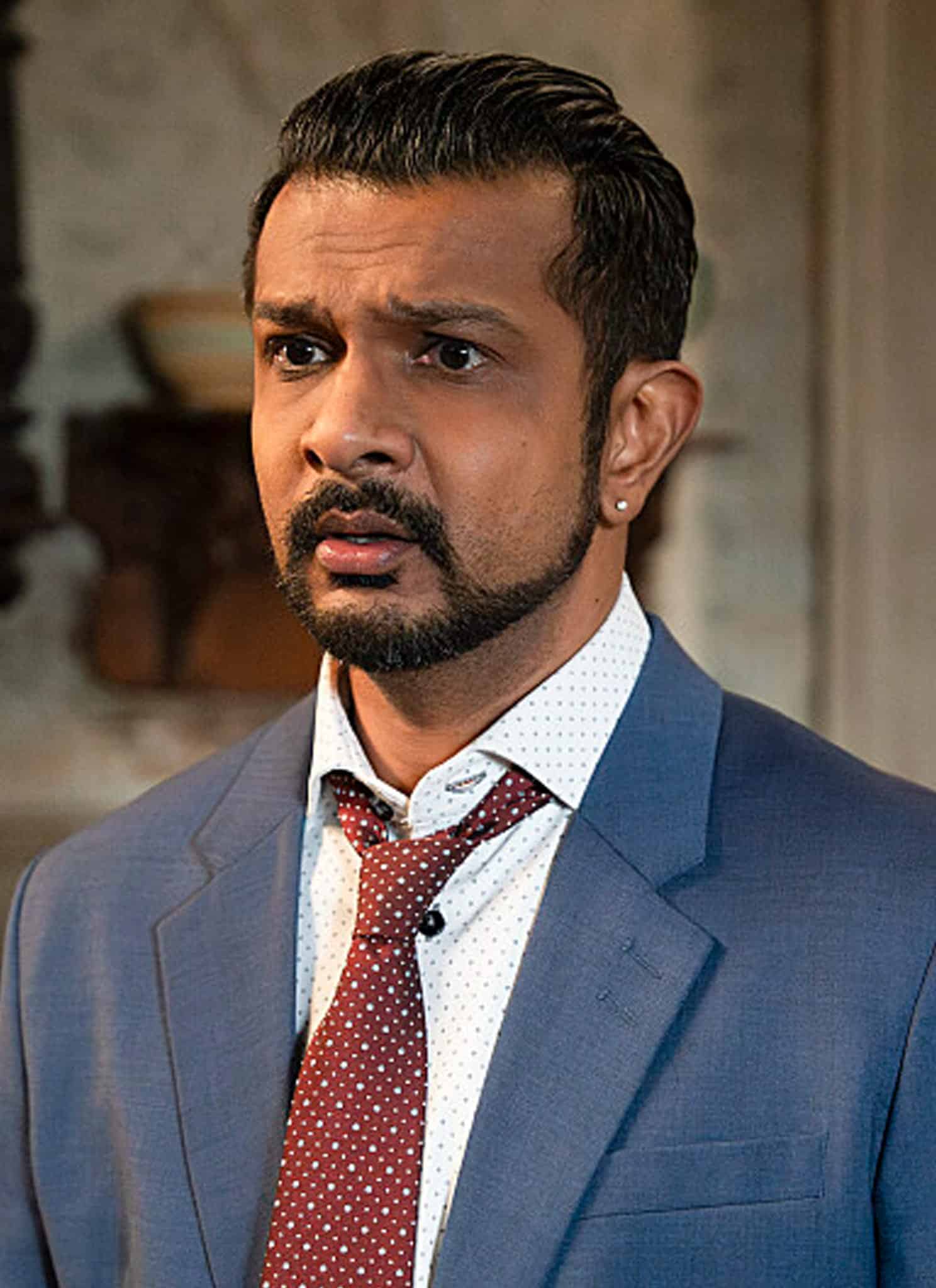 A man in a blue suit with a white shirt and red tie looks concerned, standing indoors with blurred warm lights and a chandelier in the background.